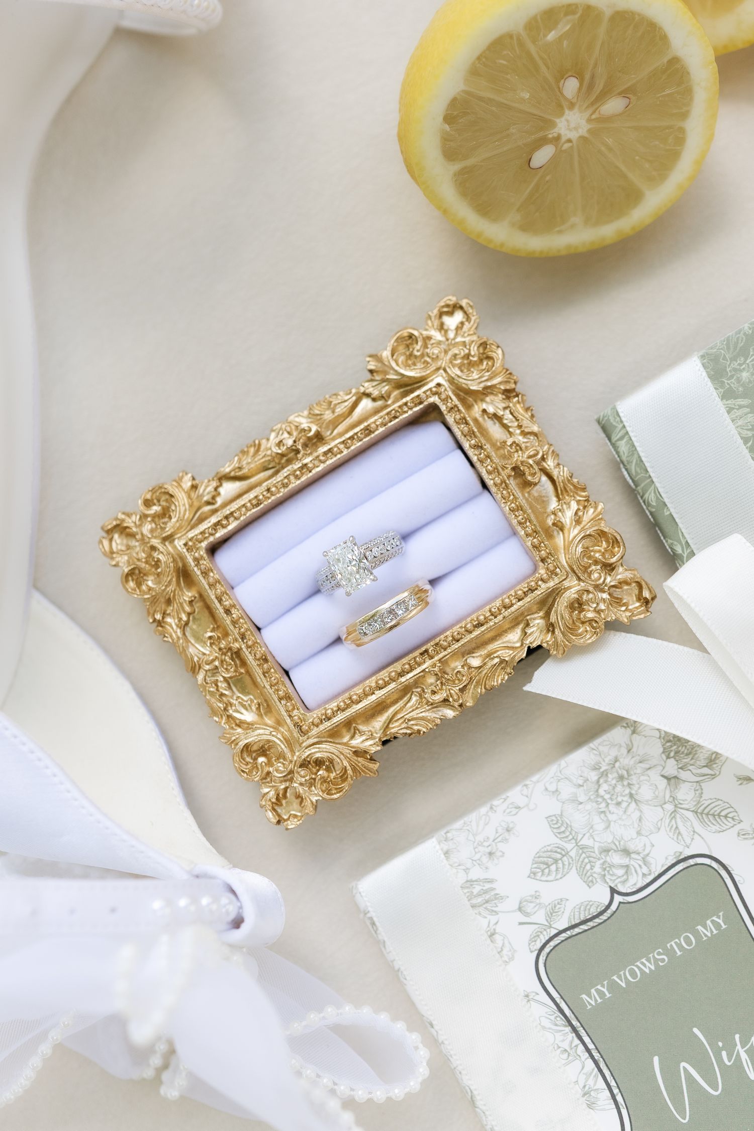 Diamond wedding rings displayed in ornate gold frame ring box with lemon and vow book nearby.