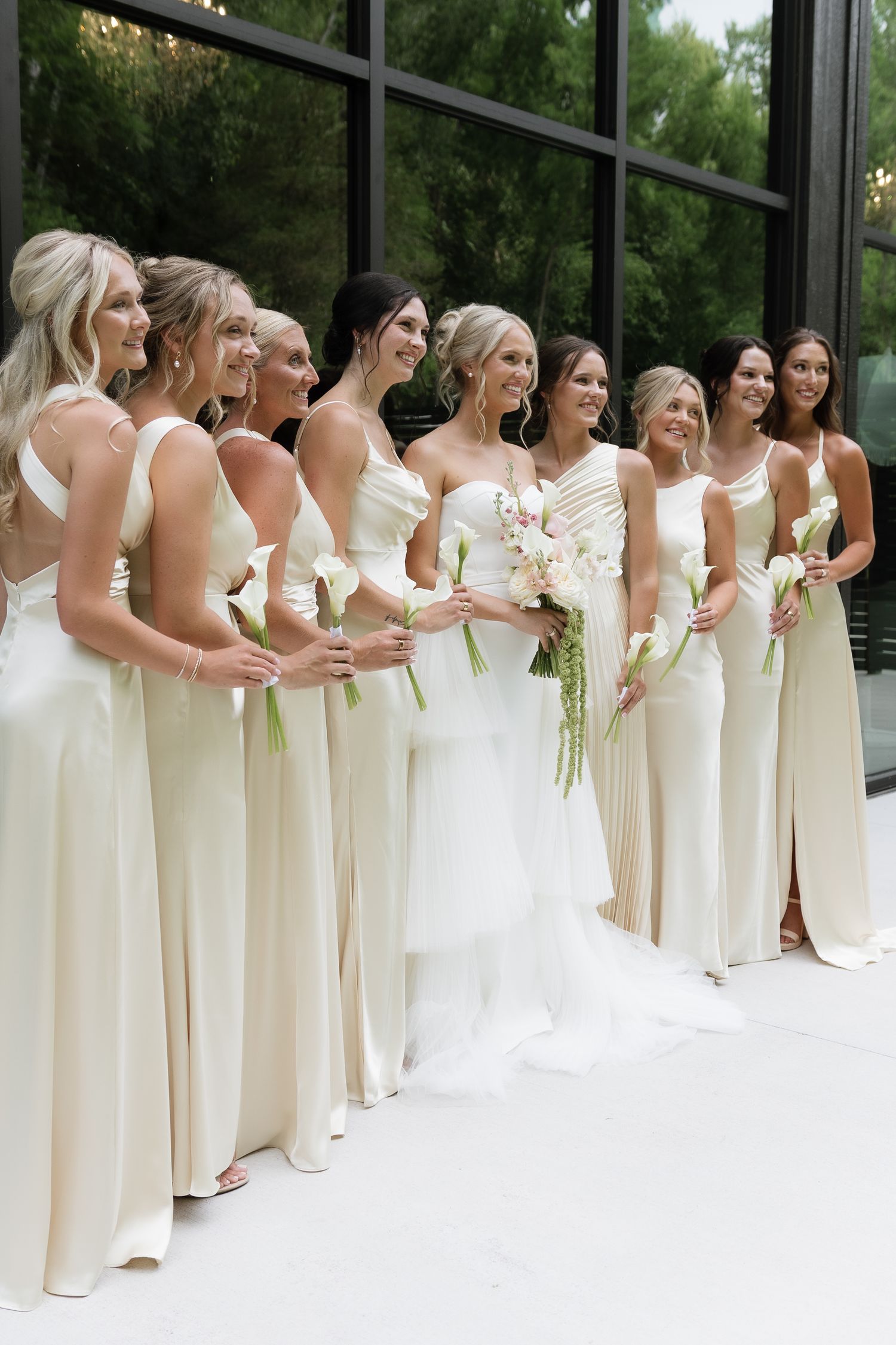 Bride with bridesmaids in ivory gowns holding white calla lilies outside a modern glass venue.