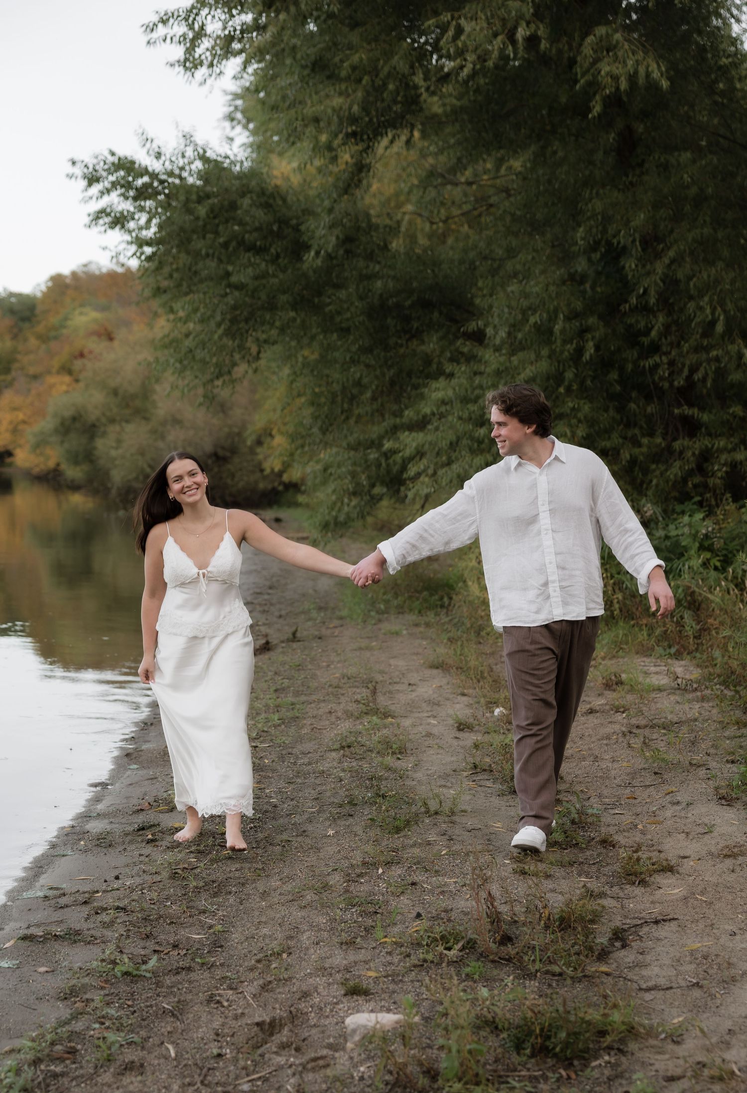 Joyful couple holding hands and walking barefoot along a lakeside shoreline with fall trees in background.
