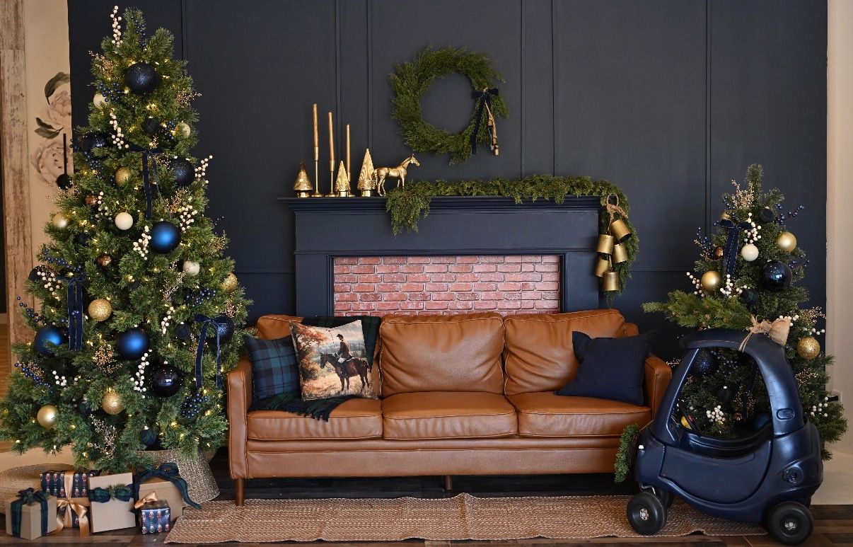 Festive living room with leather sofa, two decorated Christmas trees, wreath above brick fireplace, and blue toy car.