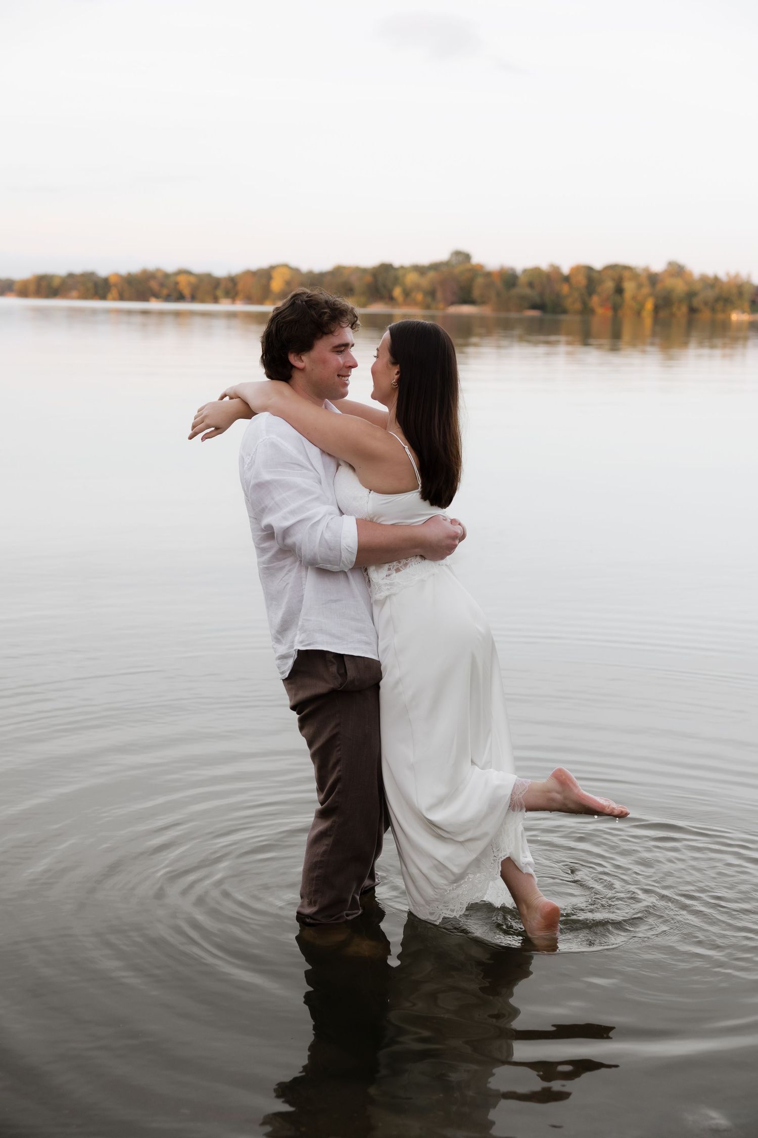 A romantic couple embraces in a lake, woman in white dress lifted by man in white shirt, autumn trees reflected in calm water.