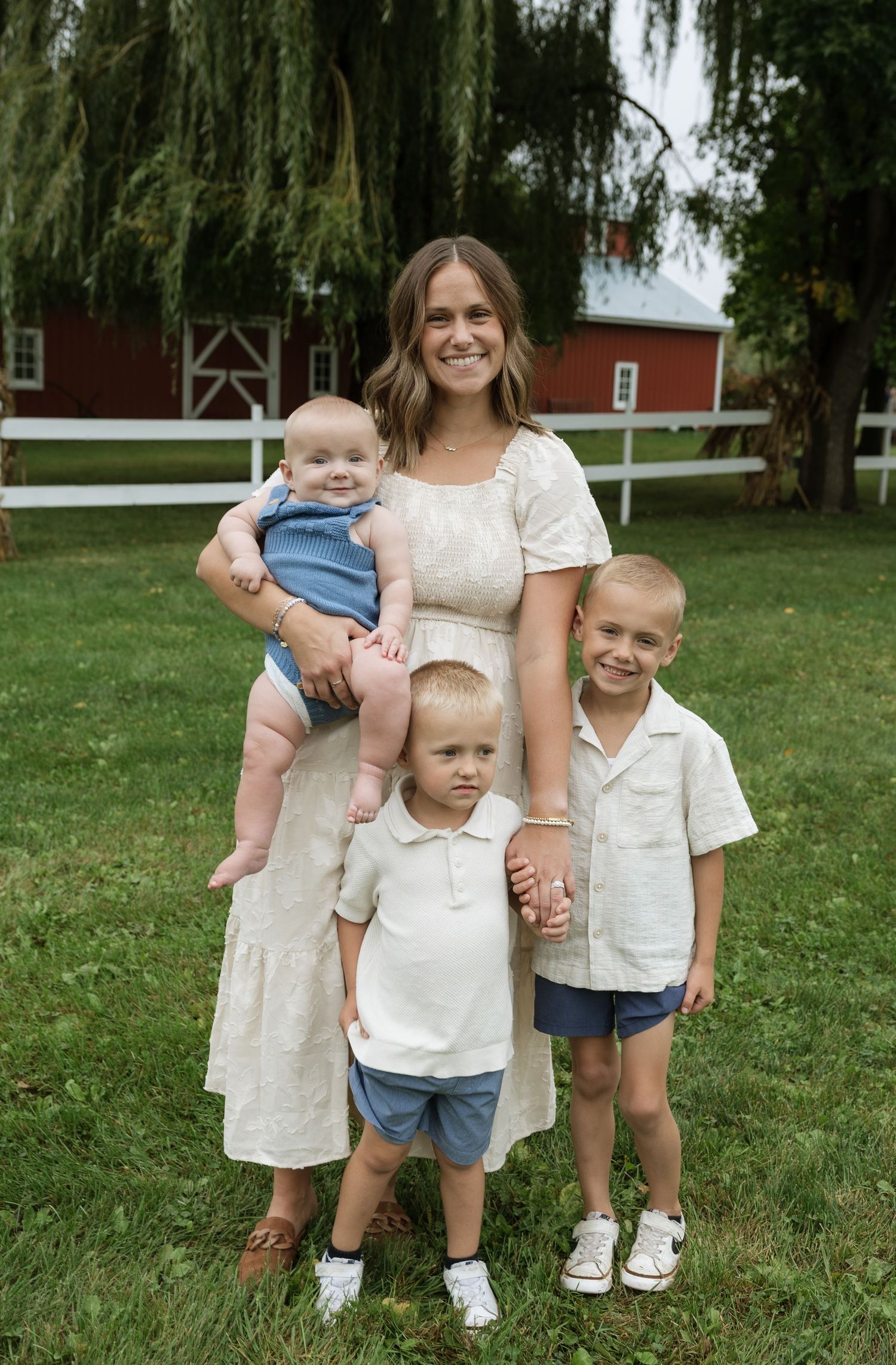 A smiling woman in a white dress holds a baby while posing with two young boys outdoors near a red barn.