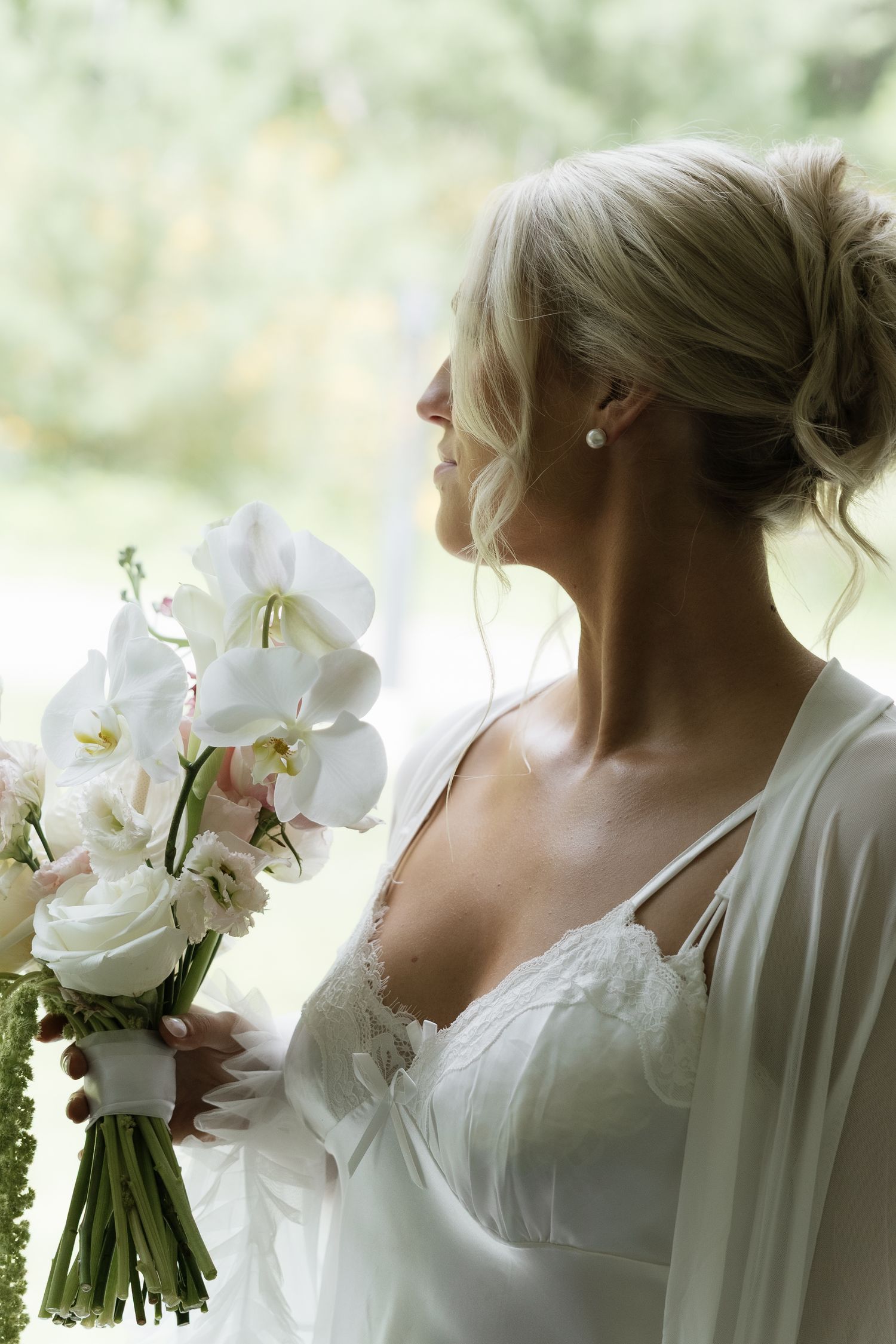Bride in white lace gown holding elegant white orchid bouquet, standing by window in soft natural light.