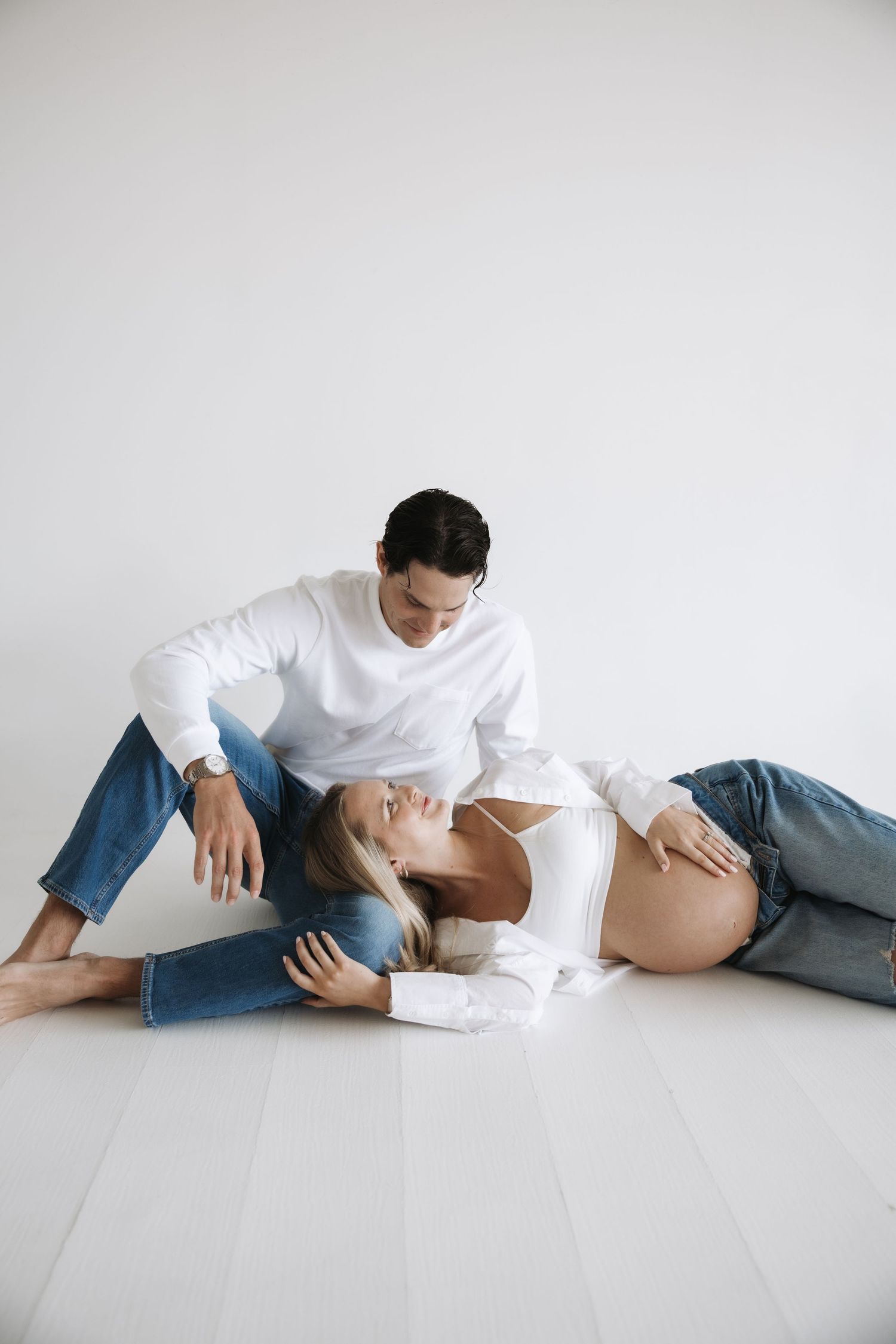 Maternity photo of pregnant couple on white floor, woman lying back showing bump, man seated beside her.