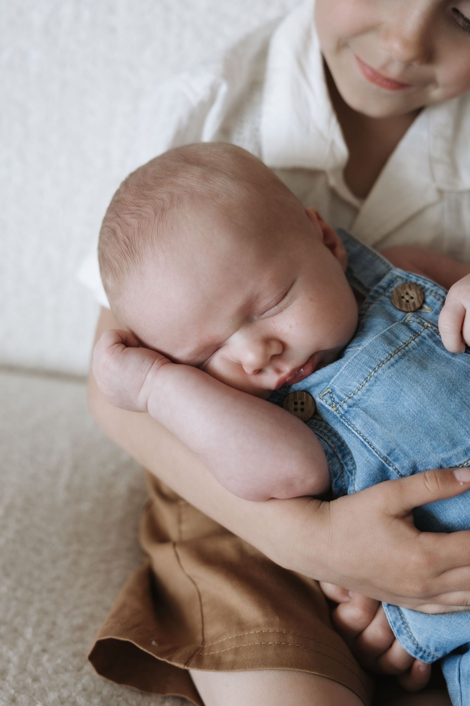 Smiling older child gently cradles sleeping newborn baby in a blue denim romper in their arms.