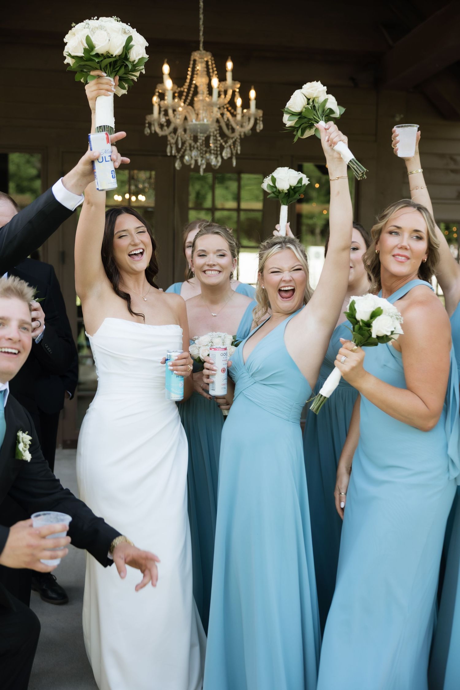 Joyful bride in white strapless gown celebrates with bridesmaids in blue dresses, raising bouquets under chandelier.