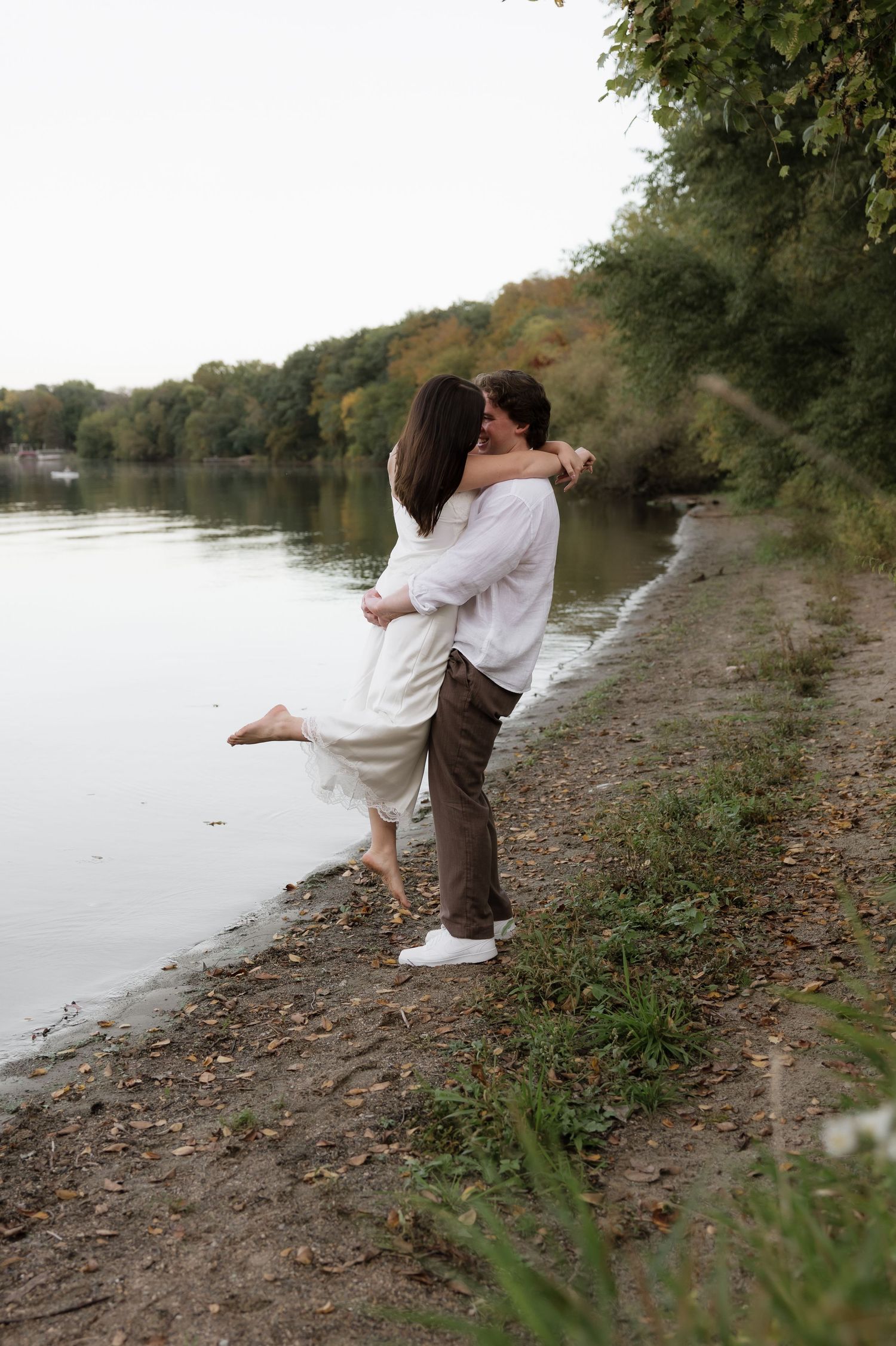 Couple embracing near a lakeside path, woman in white dress being lifted by man in white linen shirt.