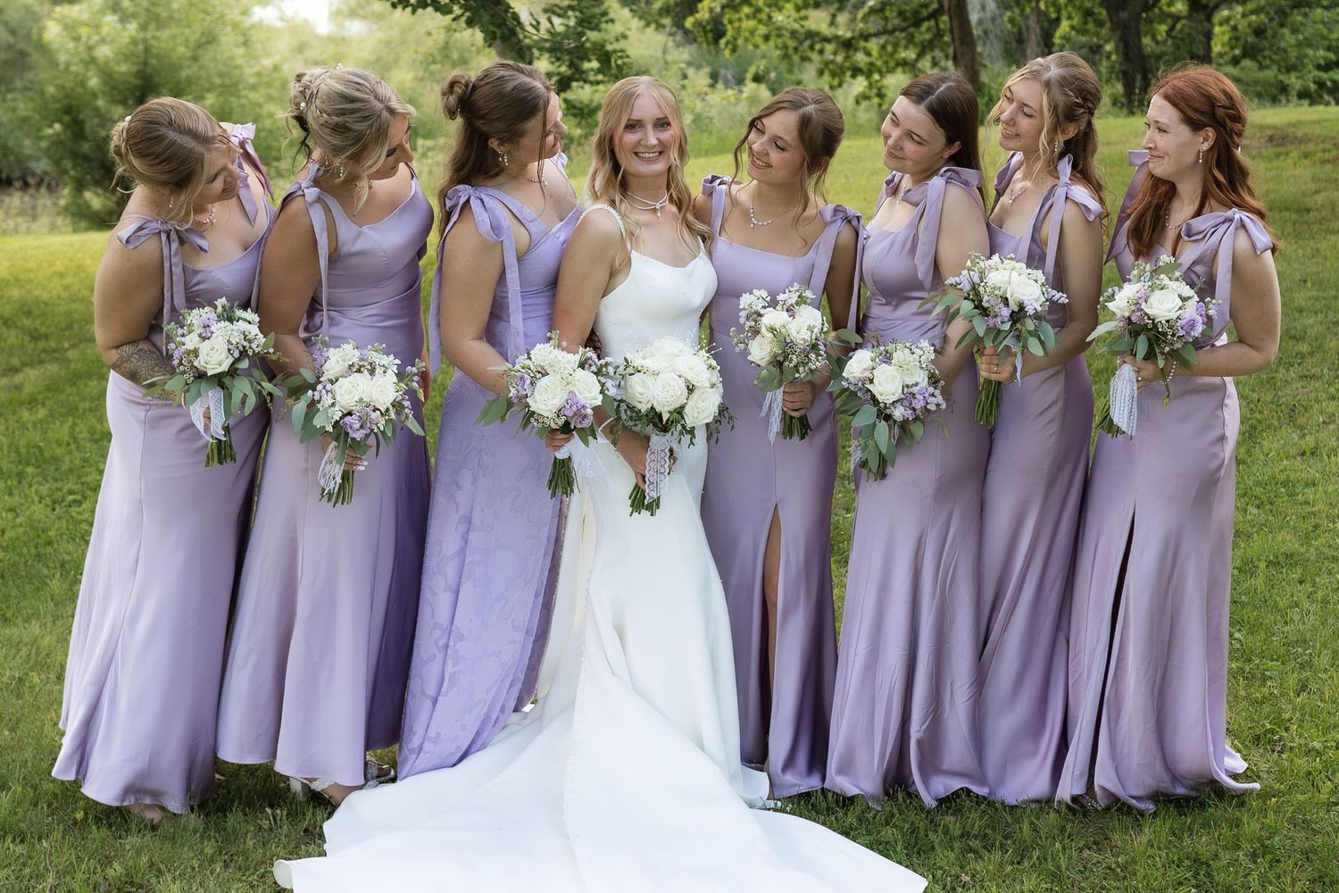 Bride in white gown surrounded by seven bridesmaids in lavender dresses holding white bouquets outdoors.