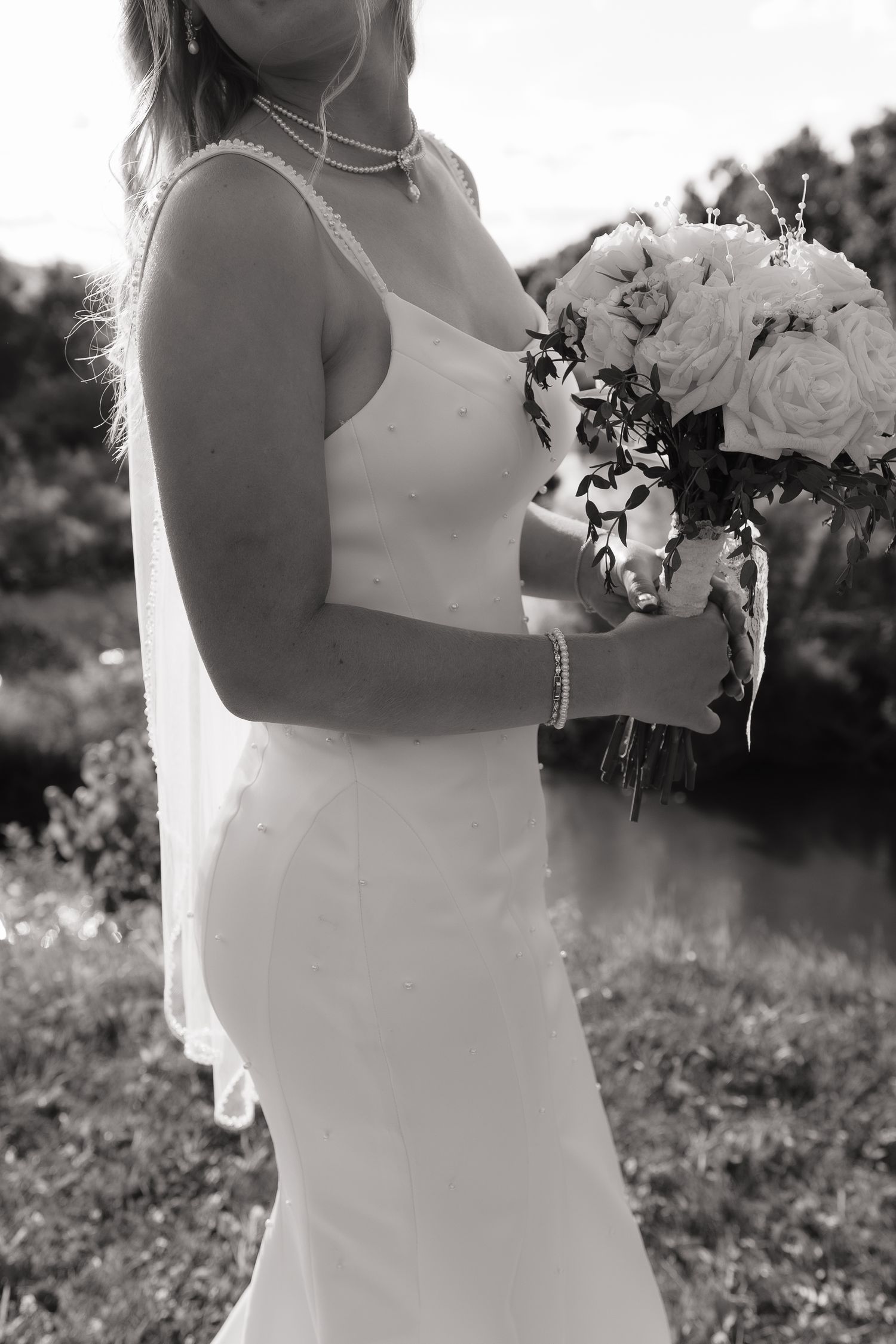 Black and white photo of a bride in a white gown holding a rose bouquet outdoors near a pond.