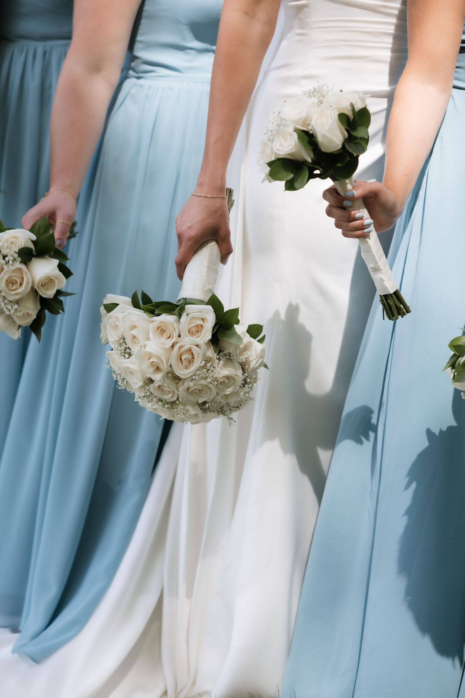 Close-up of bride and bridesmaids holding elegant white rose bouquets in white and light blue gowns.