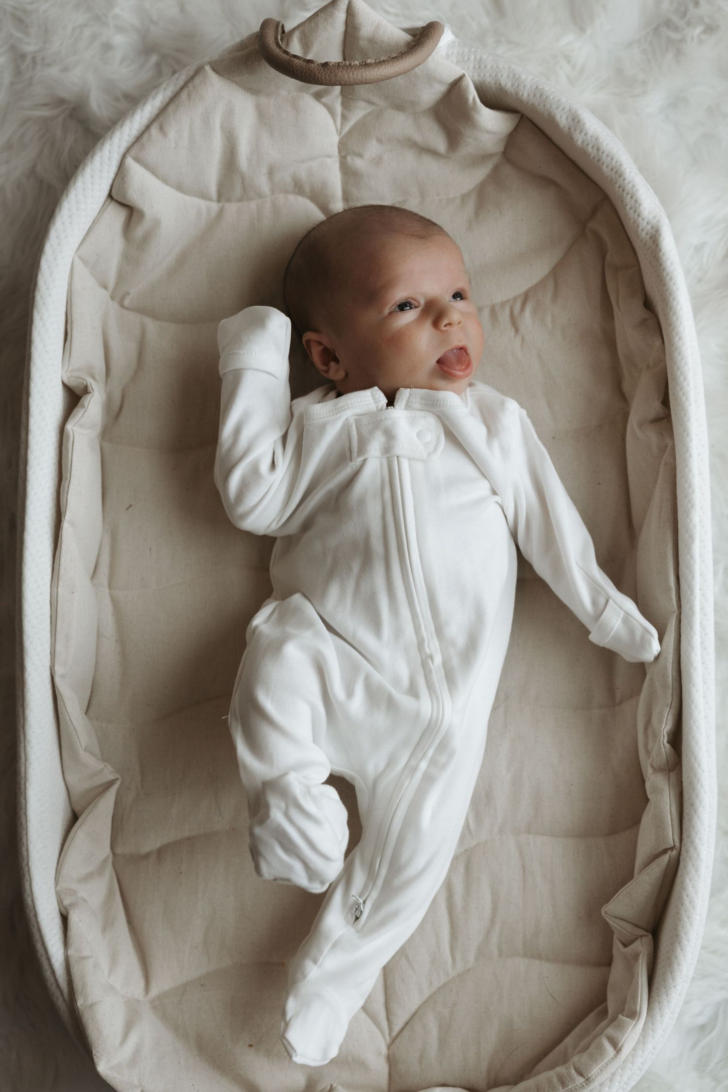 Newborn baby in a white zip-up sleepsuit lying in a beige Moses basket.