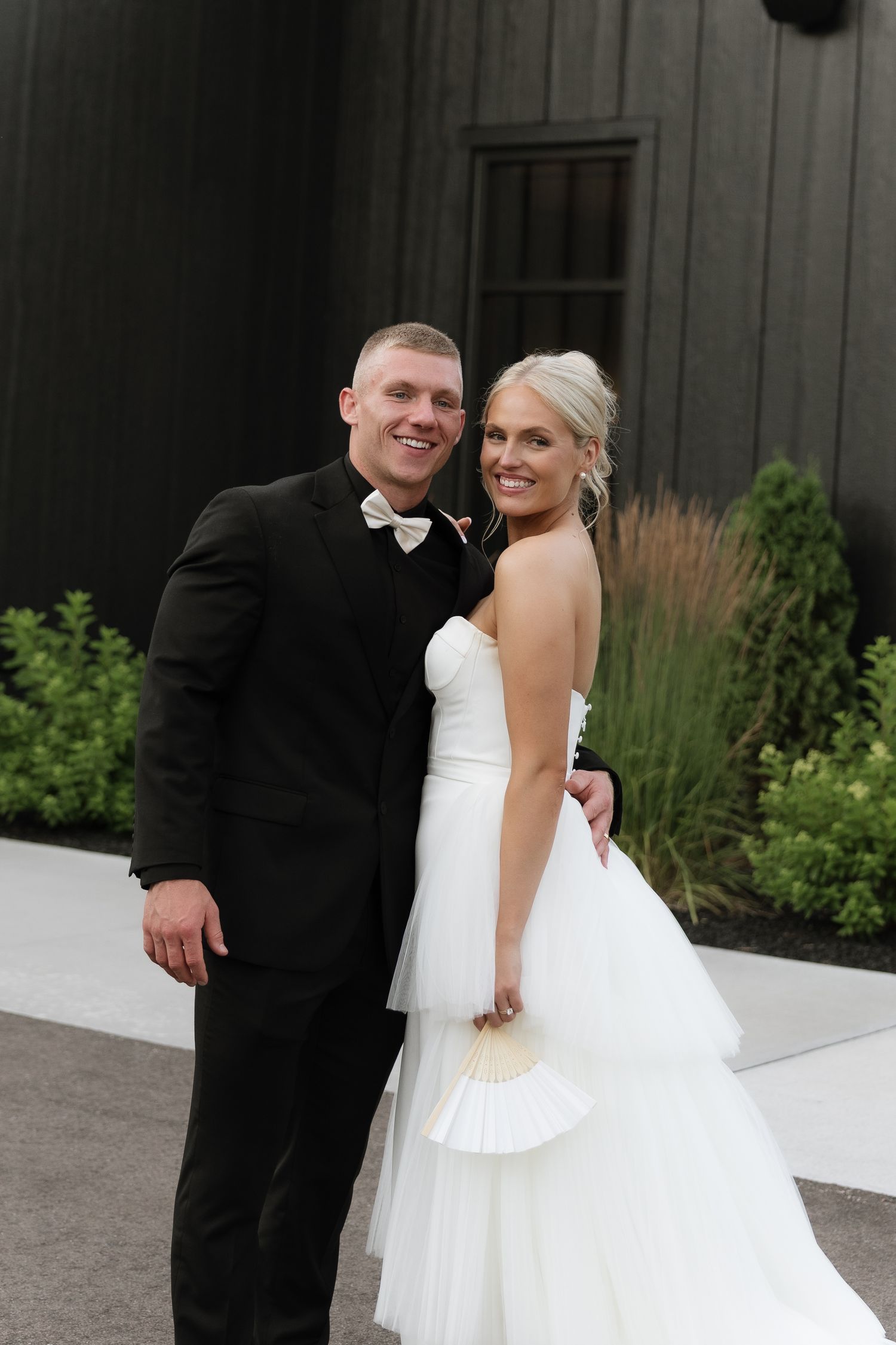 Smiling bride and groom posing together outside a dark barn, groom in black tuxedo with bow tie.