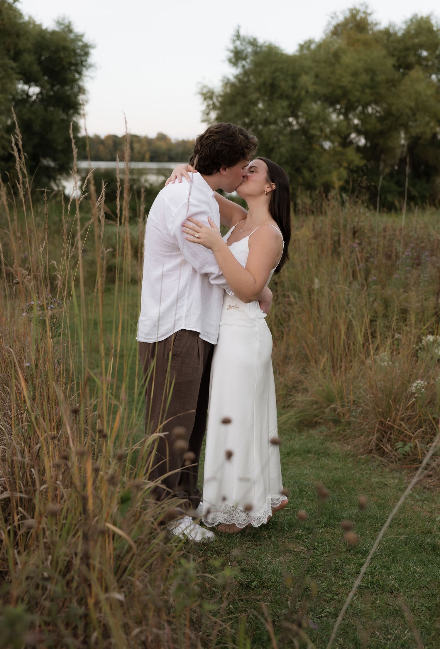 Couple sharing a romantic kiss in a golden meadow field near a lake during fall engagement session.