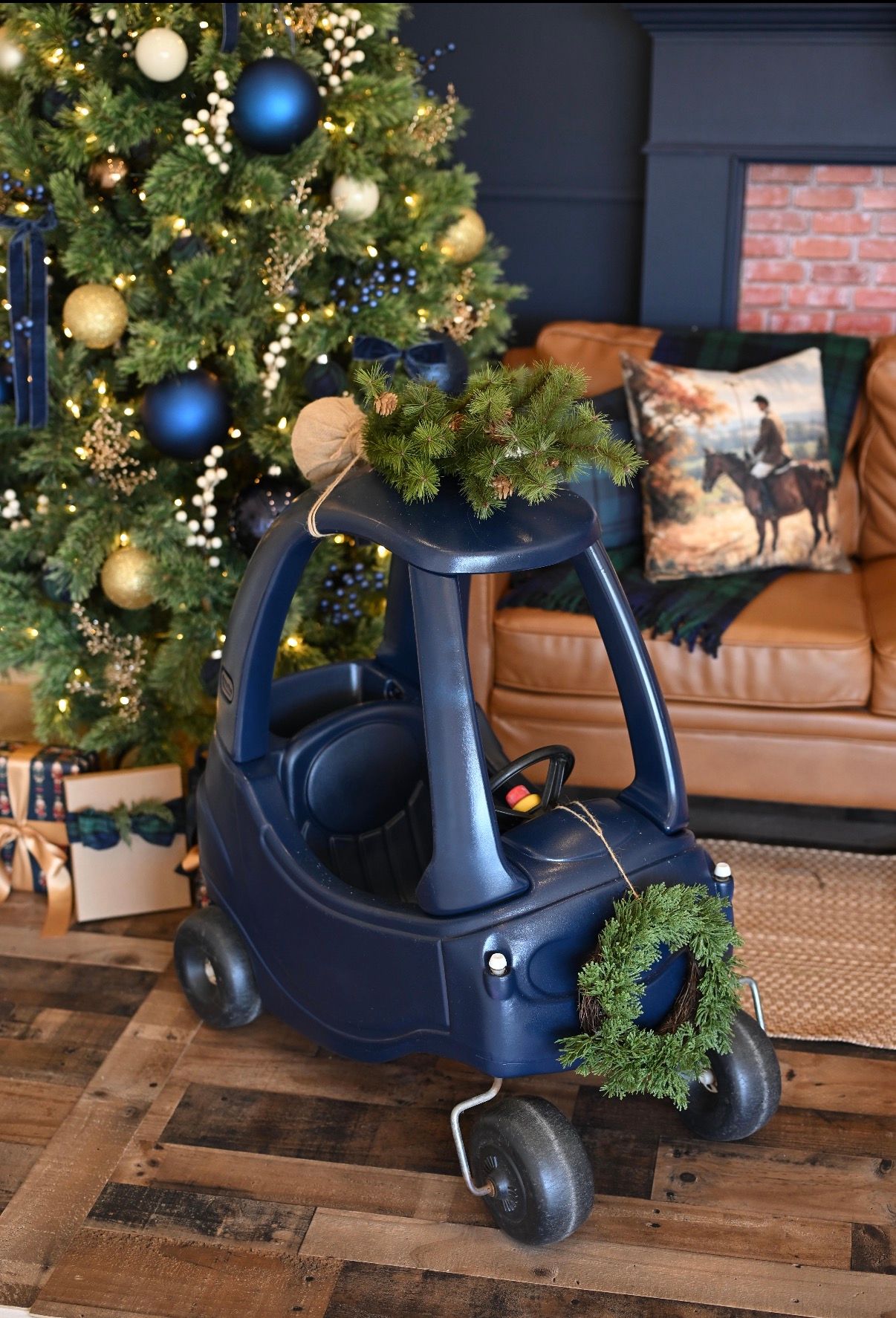Navy blue Little Tikes Cozy Coupe toy car decorated with a Christmas wreath beside a decorated holiday tree with gold and blue ornaments.