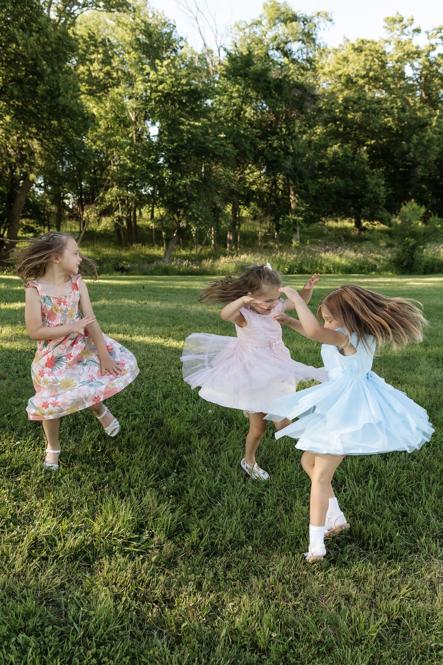 Three young girls in colorful dresses twirl and dance joyfully on green grass.
