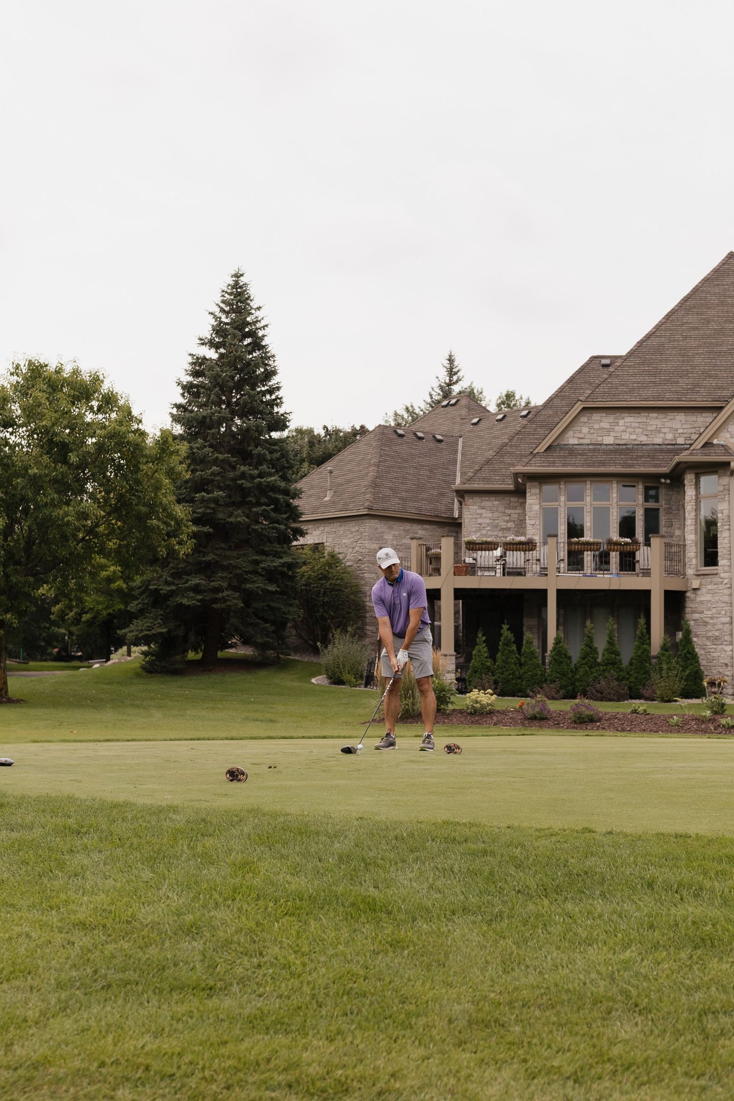 Man in purple shirt putting on a private backyard golf green beside a large brick house surrounded by trees.