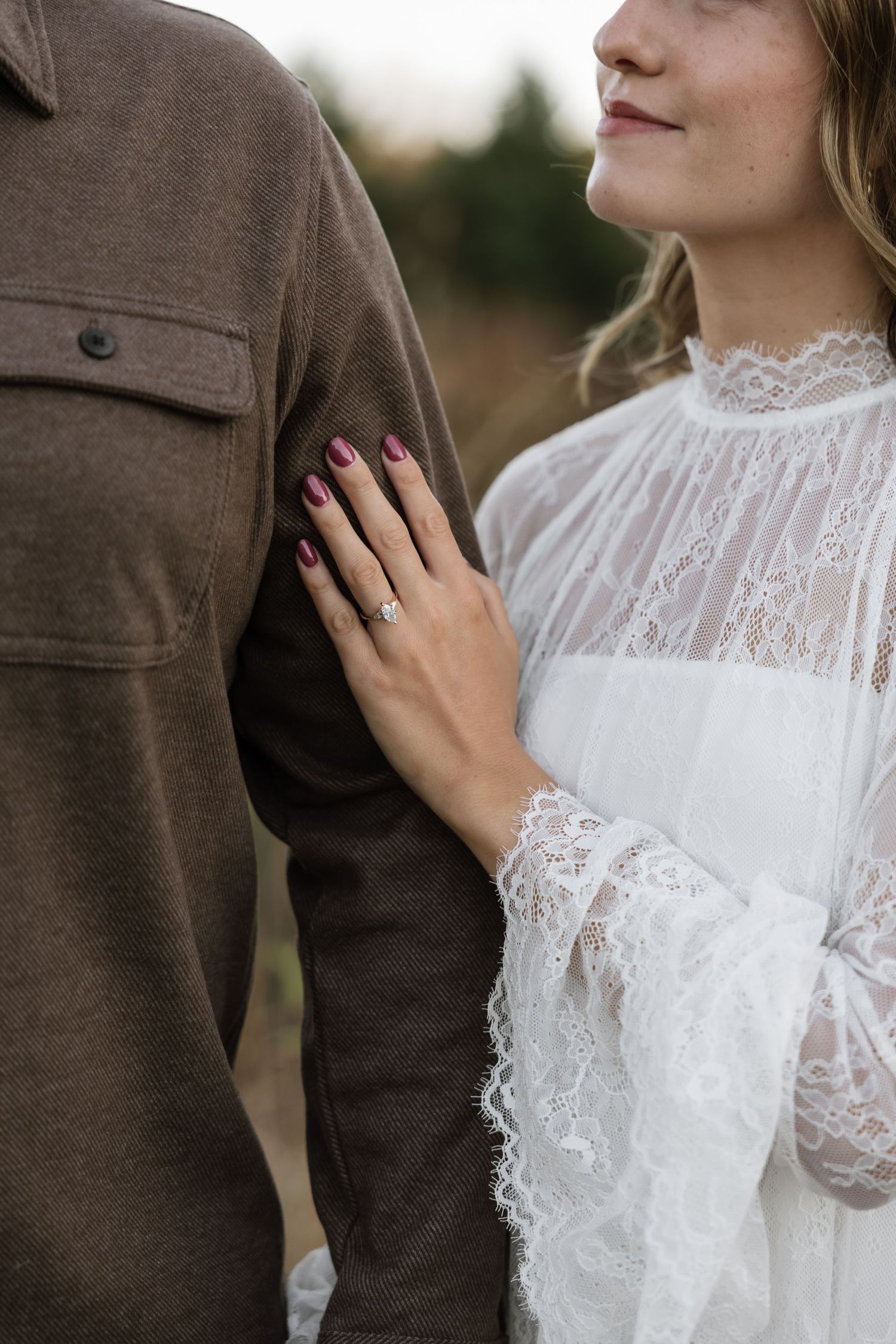 Close-up of bride in white lace dress showing diamond engagement ring with burgundy nails on groom's brown jacket.