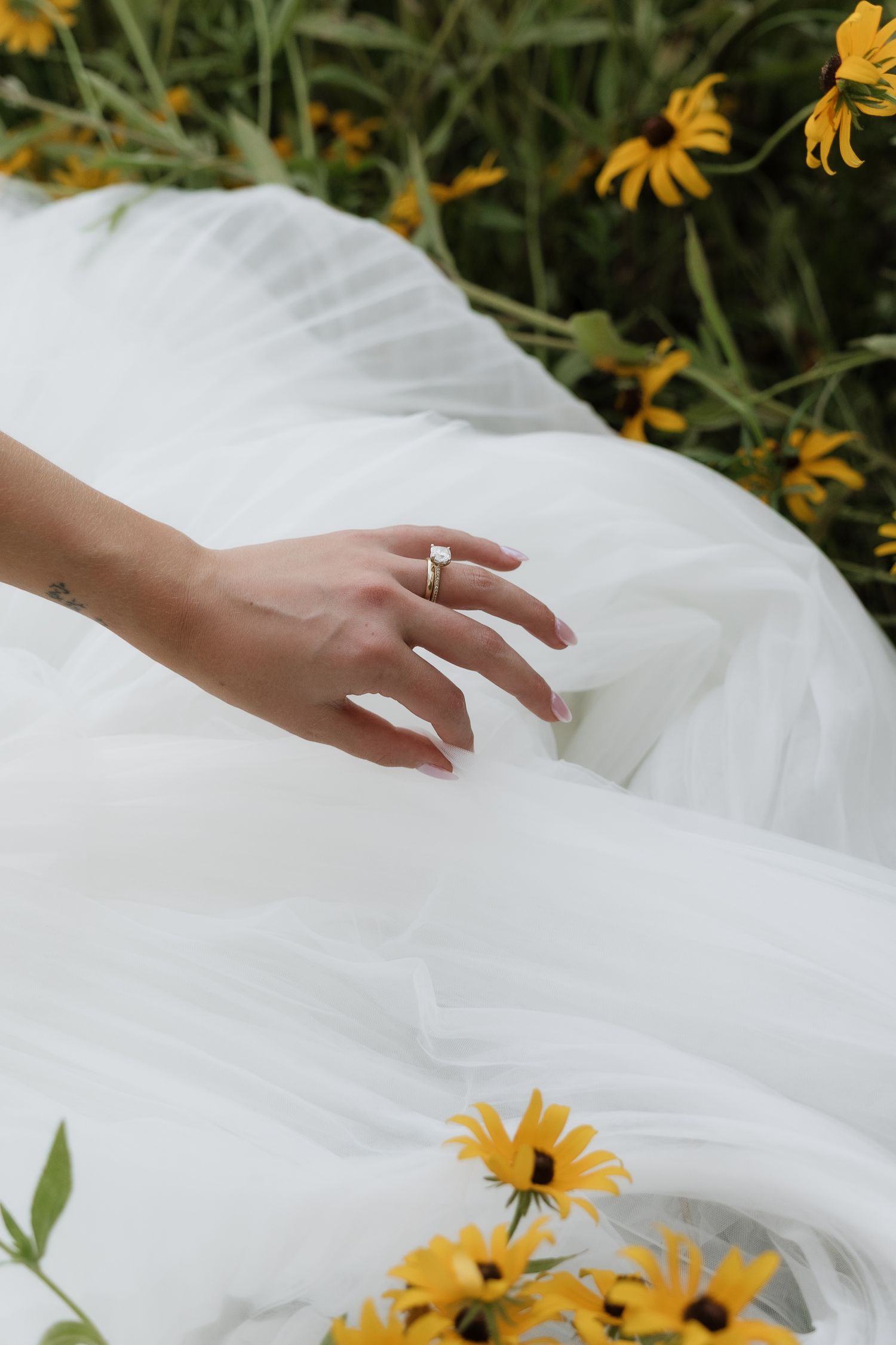 Close-up of bride's hand displaying engagement ring on white tulle gown among yellow wildflowers.