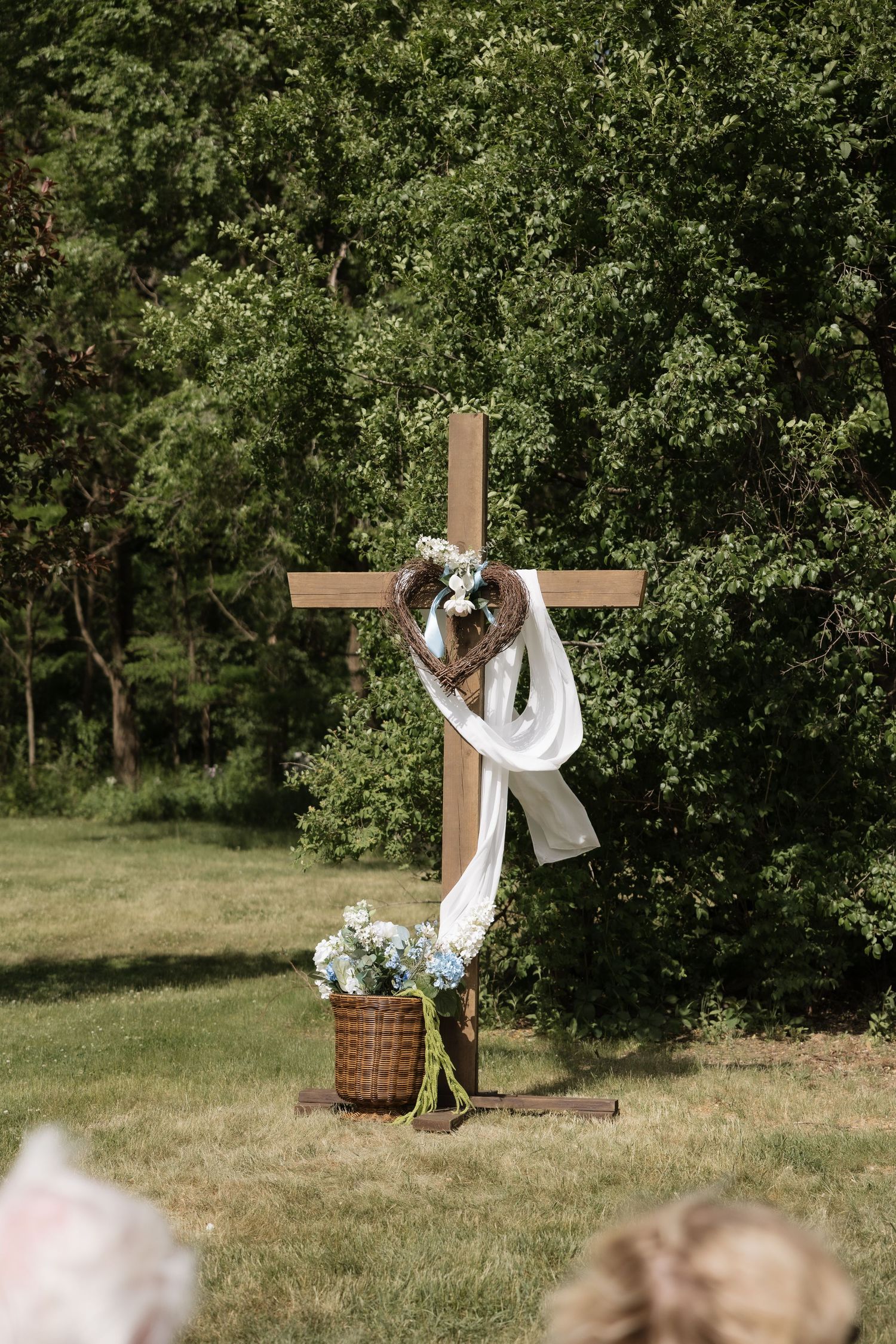 Rustic wooden cross decorated with white fabric, a heart wreath, and a basket of blue hydrangeas at an outdoor wedding.