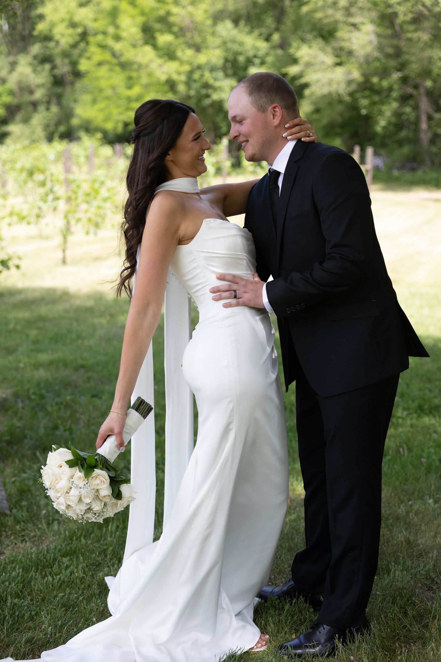 Bride and groom share romantic moment in lush green vineyard, bride in white halter gown with long train.