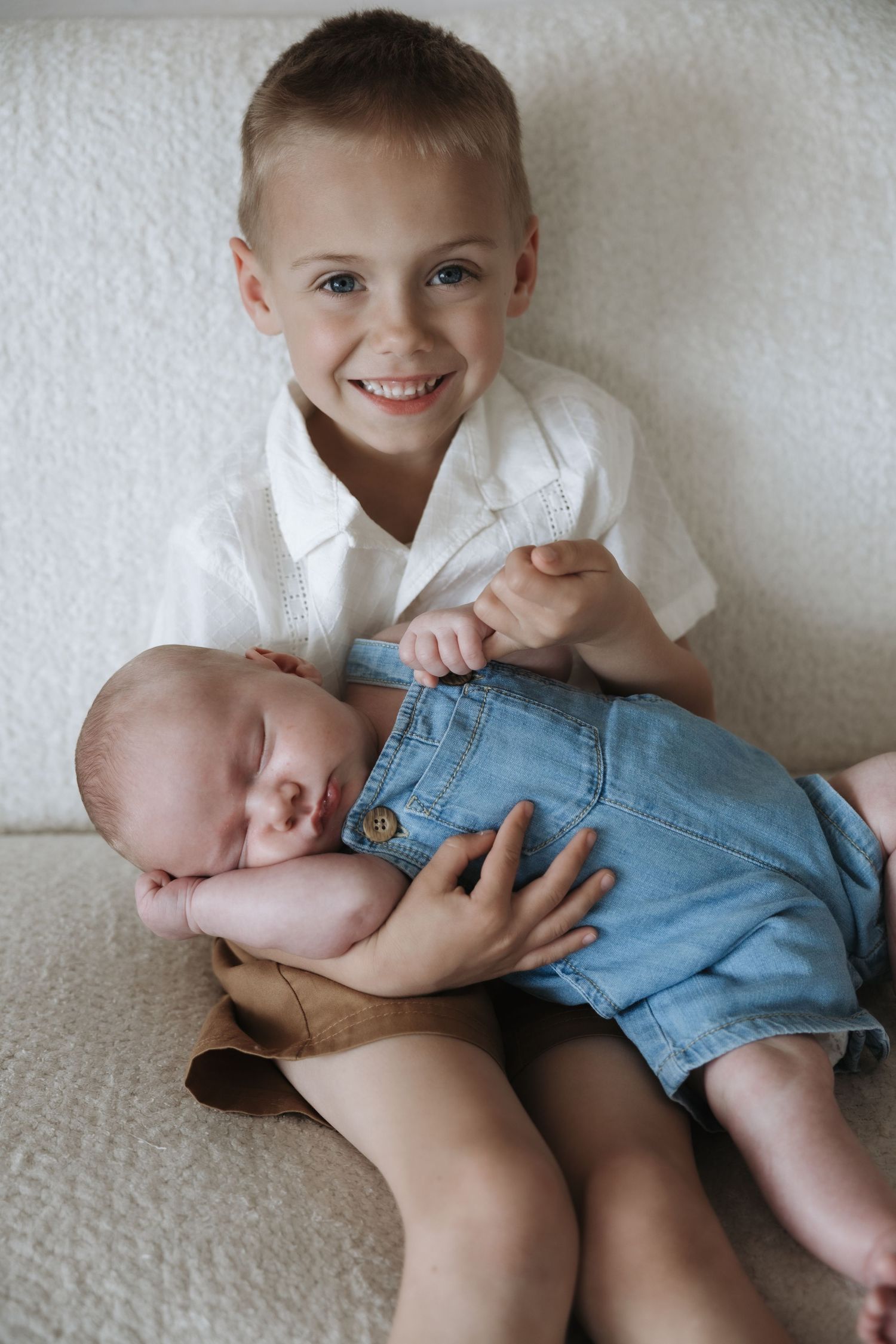Happy blue-eyed boy smiles while carefully holding sleeping newborn baby in a blue romper.