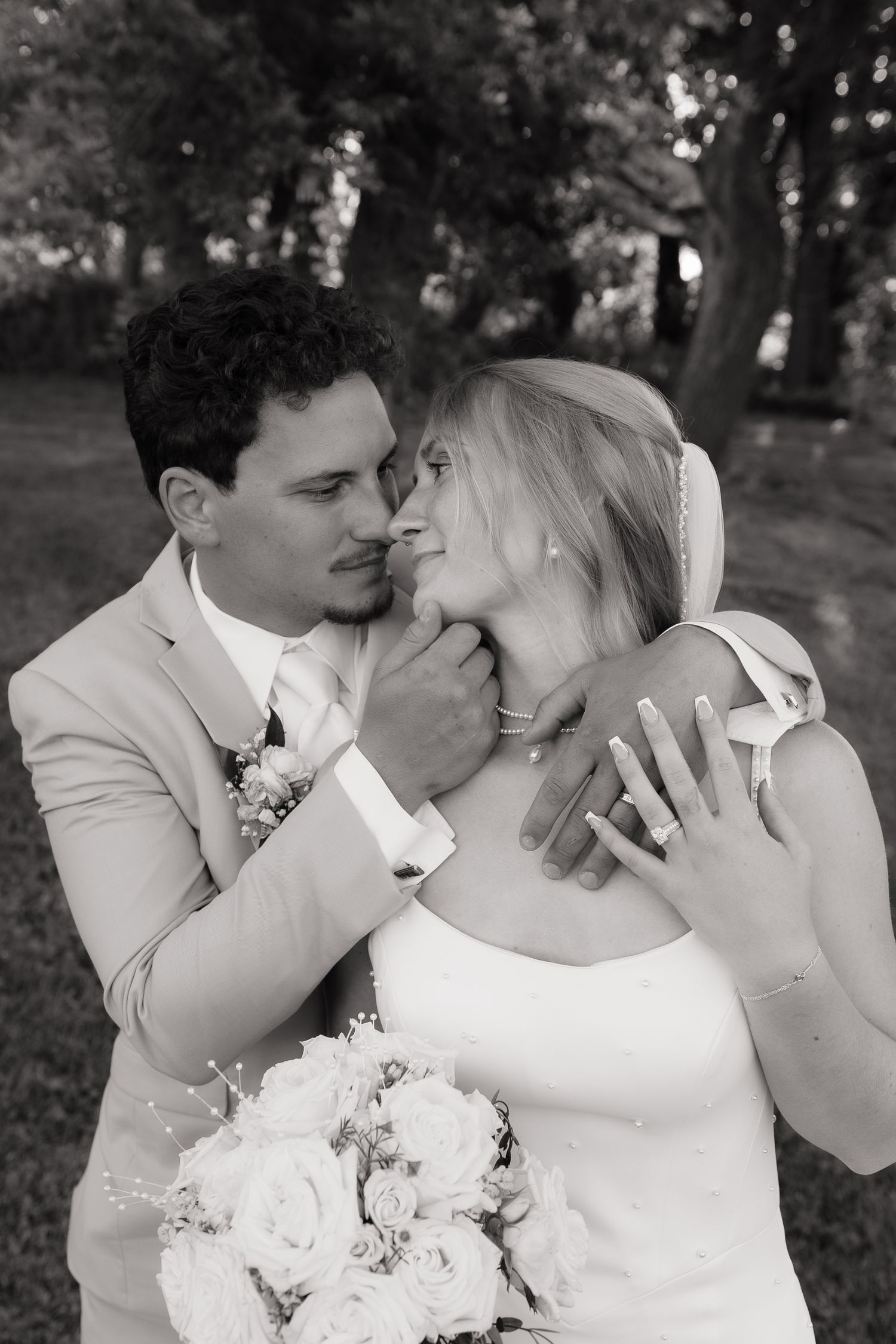 Black and white photo of a groom in a suit tenderly holding his bride in a white dress and bouquet outdoors.