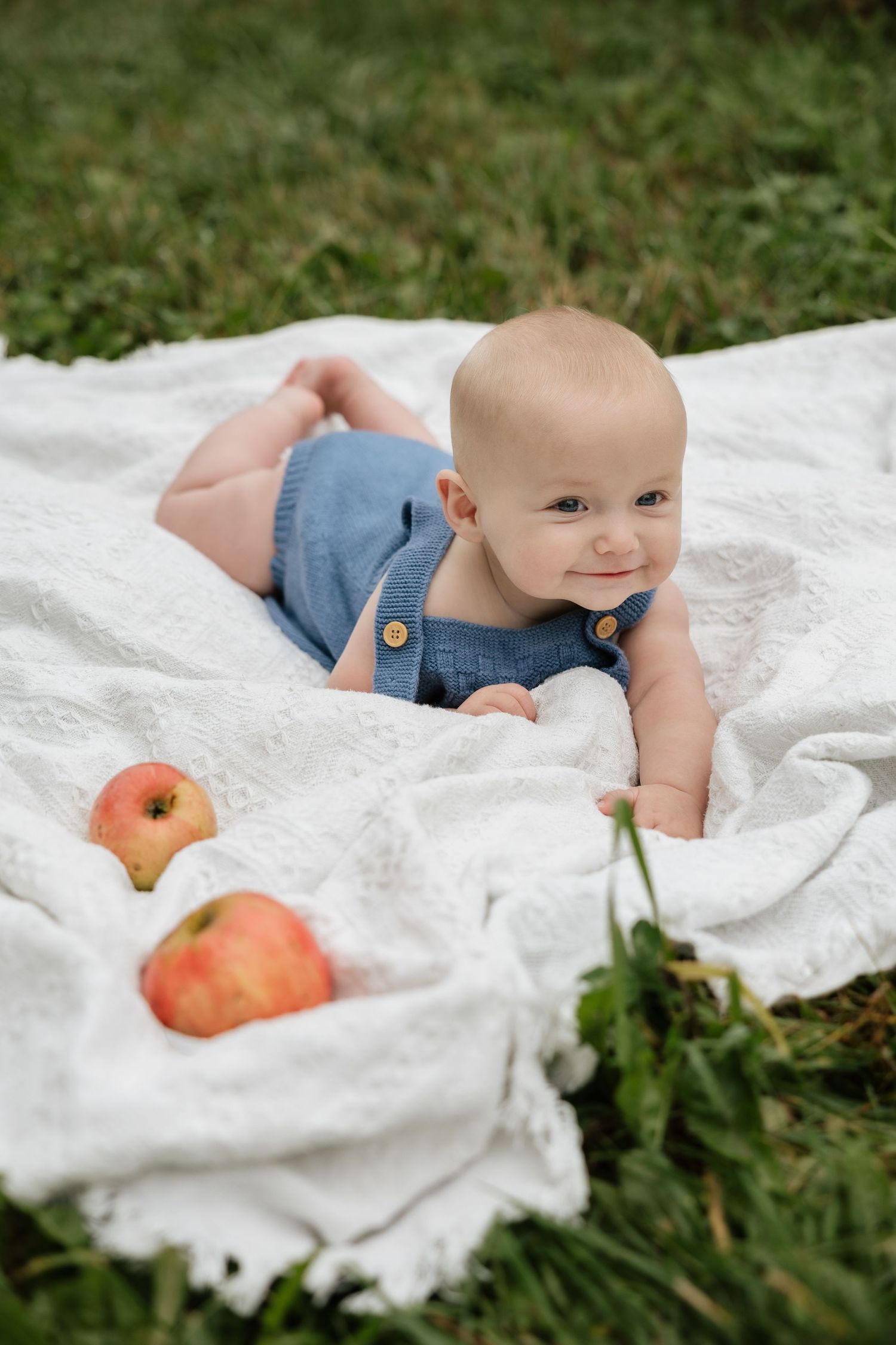 Smiling baby in blue denim overalls lying on white blanket outdoors with red apples nearby.