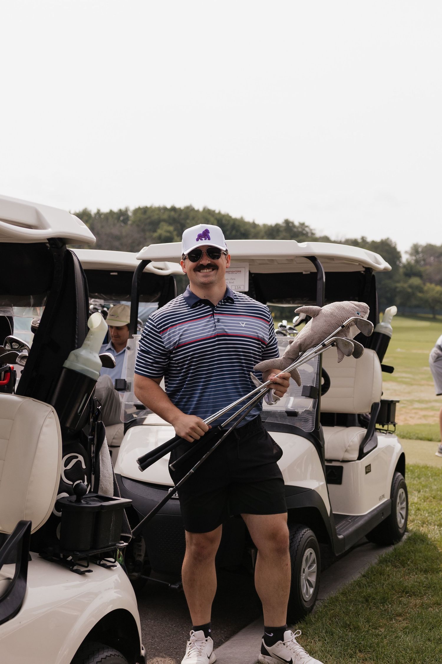 Smiling man with mustache holding golf club near cart on course, wearing striped polo and white cap.