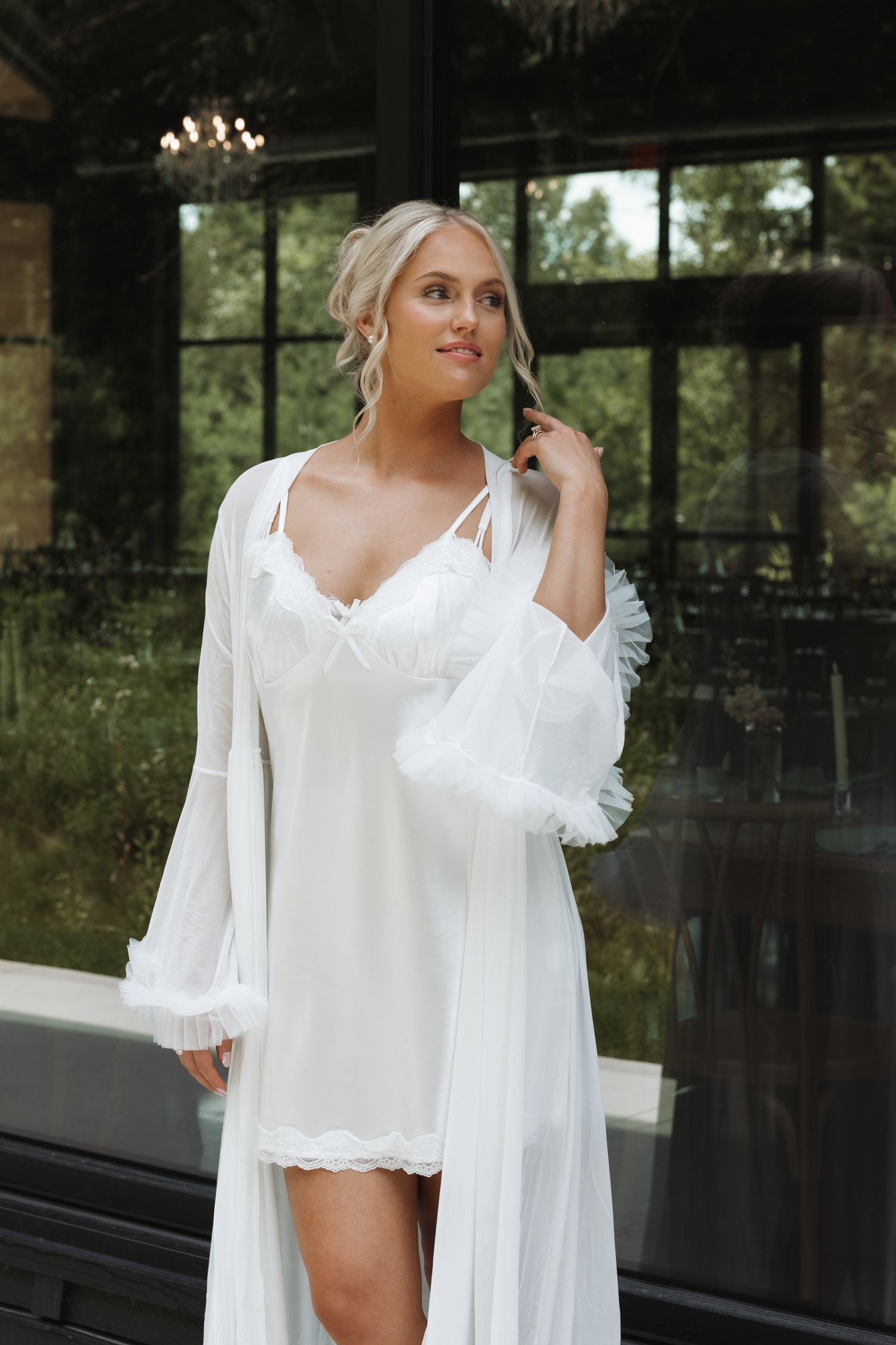 Elegant bride in white lace slip dress and flowing robe poses in luxurious indoor venue with garden views.