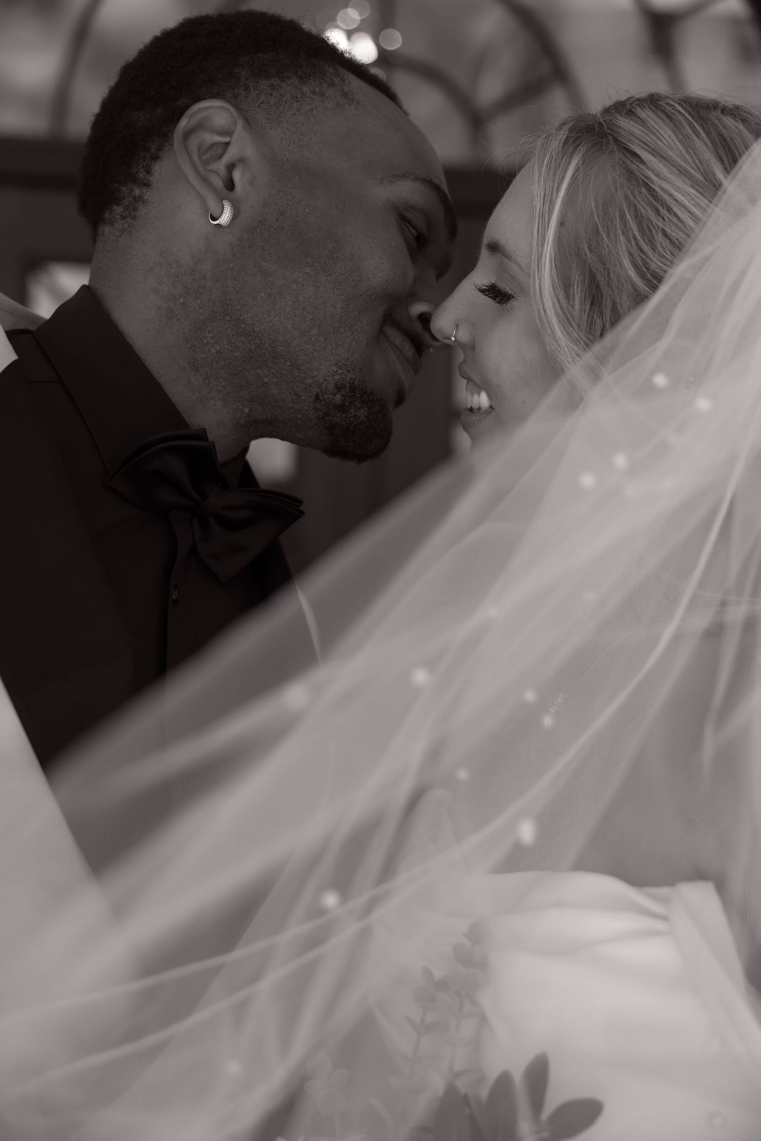 Black and white wedding photo of an interracial couple sharing a tender moment under a flowing bridal veil.