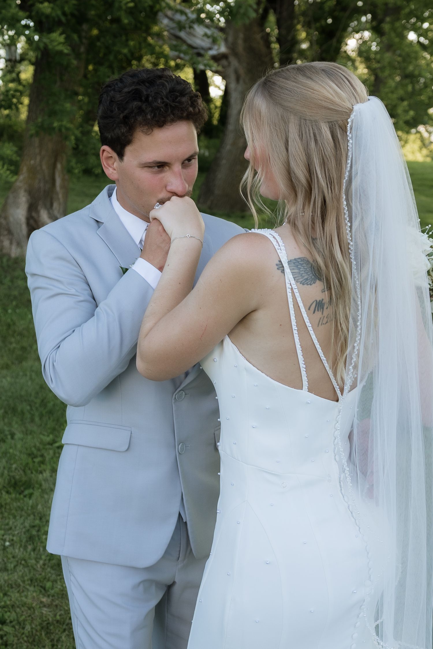 Groom in light gray suit kissing bride's hand outdoors, bride wearing white wedding dress with veil and back tattoo.