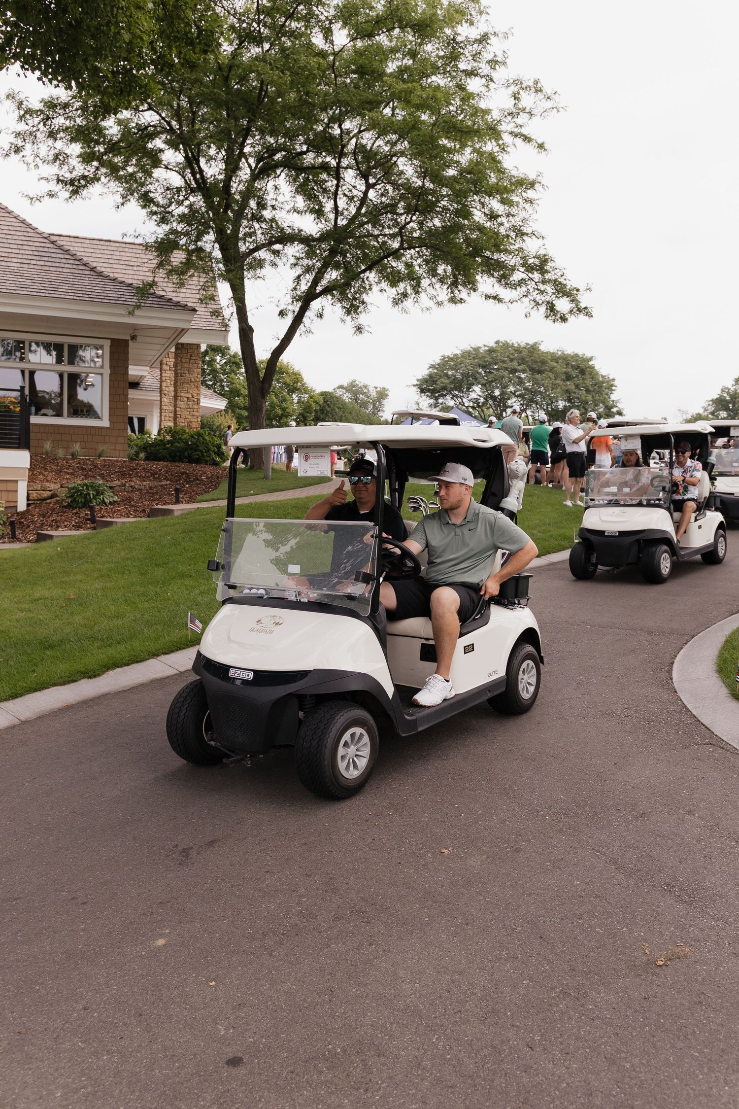 Two men ride a white golf cart along a paved path at a golf course, with more carts visible behind them.