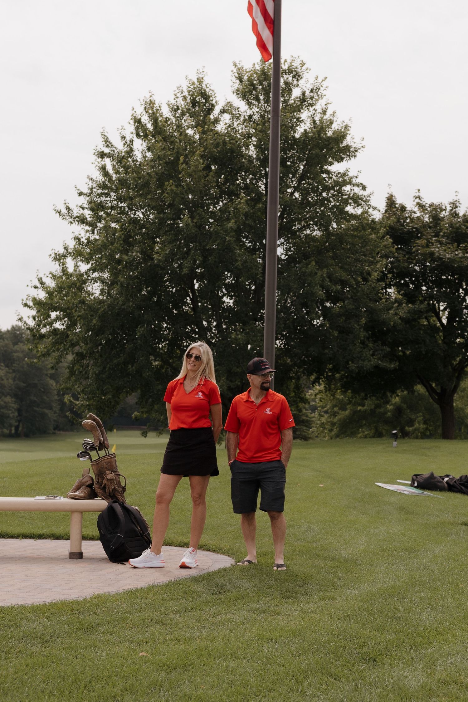 Two golfers in matching red polo shirts stand near a flagpole on a lush green golf course.
