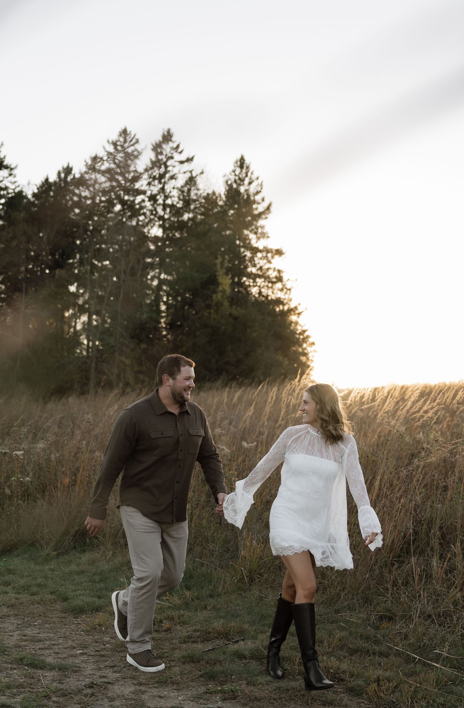 Couple holding hands and laughing while walking through a golden field at sunset, woman in white dress and boots.