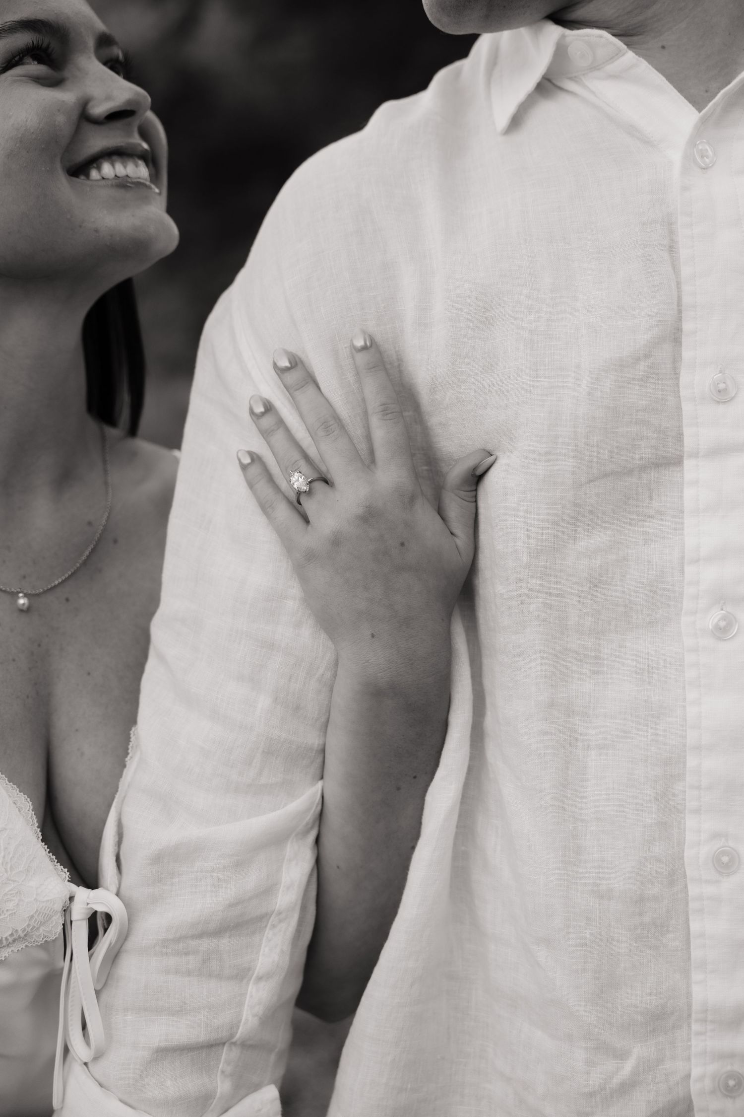 Black and white photo of a smiling woman placing her hand on a mans chest, displaying her diamond engagement ring.