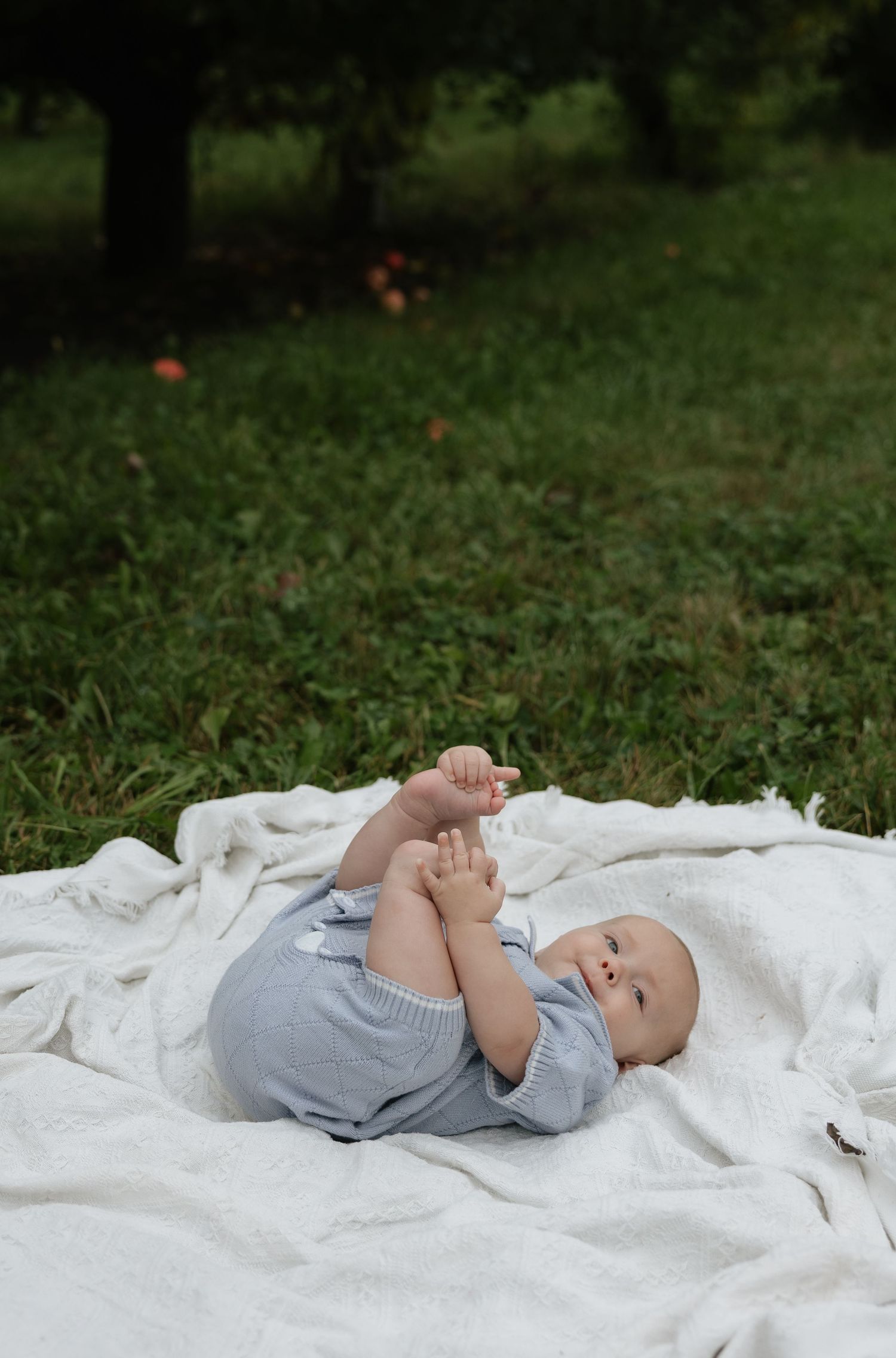 A baby in a gray romper lies on a white blanket outdoors, playfully holding their feet up in a lush green garden.