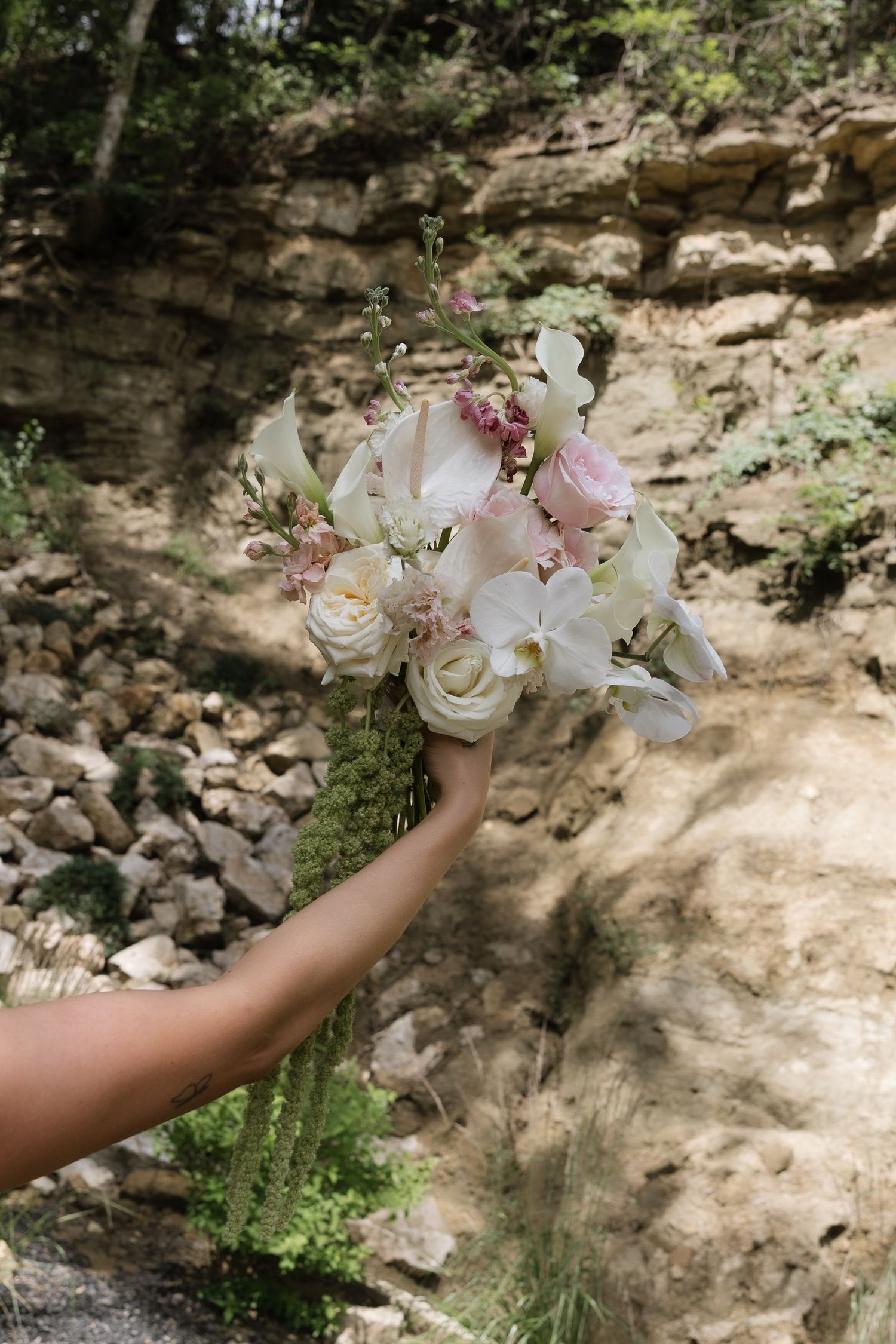 Bride holding lush white and pink wedding bouquet with orchids and roses against rocky natural backdrop.