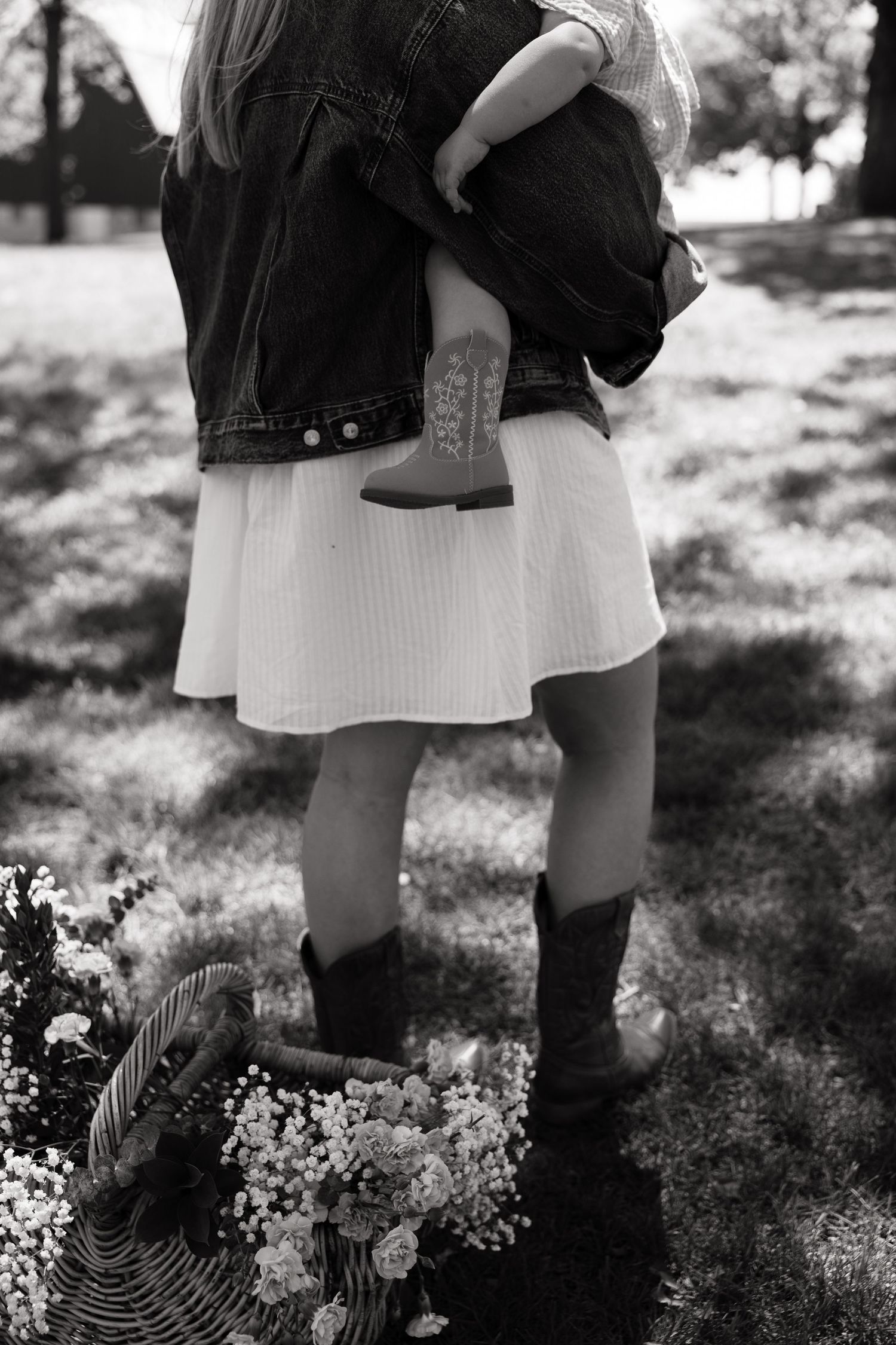 Black and white image of mother holding toddler in cowboy boots beside a flower basket outdoors.