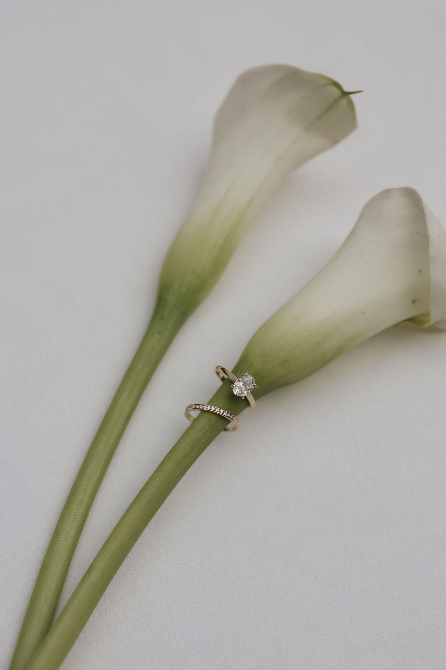 Diamond engagement ring resting on white calla lily stems against a soft gray background.