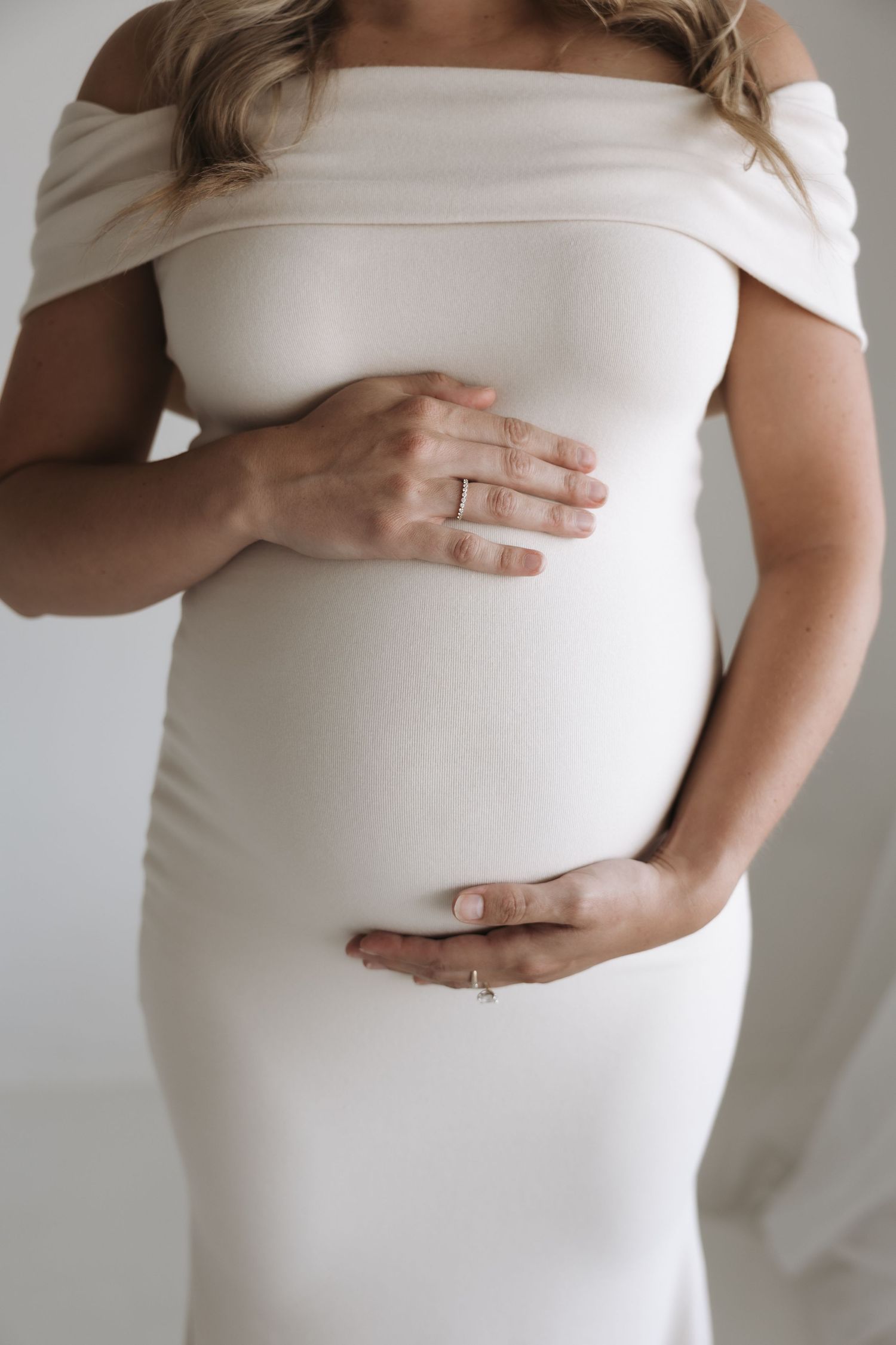 Close-up of pregnant woman in white off-shoulder dress holding her baby bump with both hands.