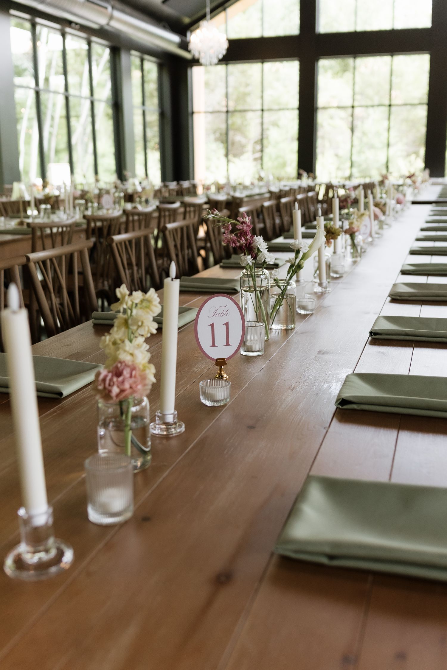 Elegant wedding reception tables set with green napkins, candles, and small floral arrangements in a glass-walled venue.