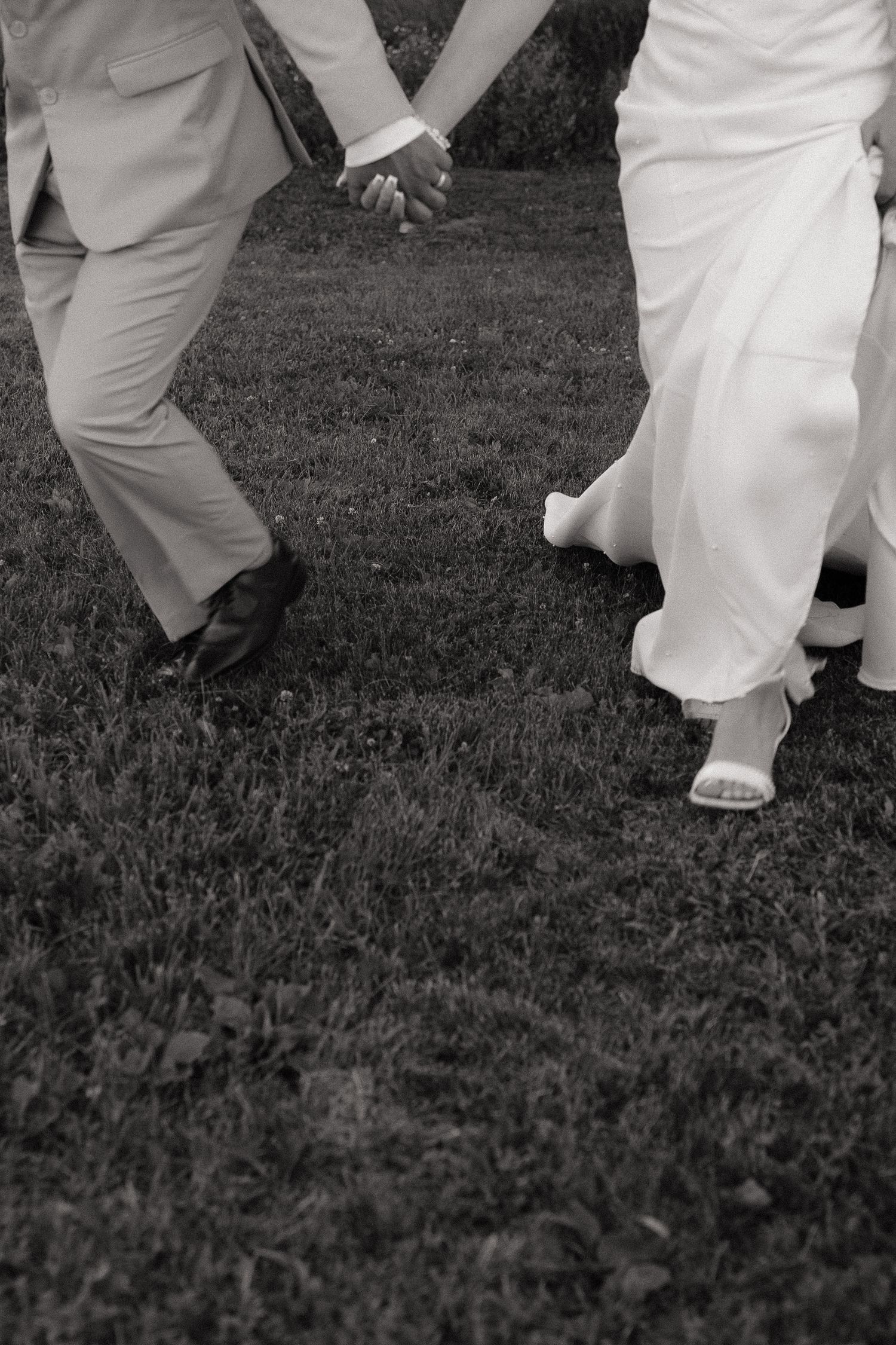 Black and white photo of a bride and groom holding hands while walking across a green lawn.
