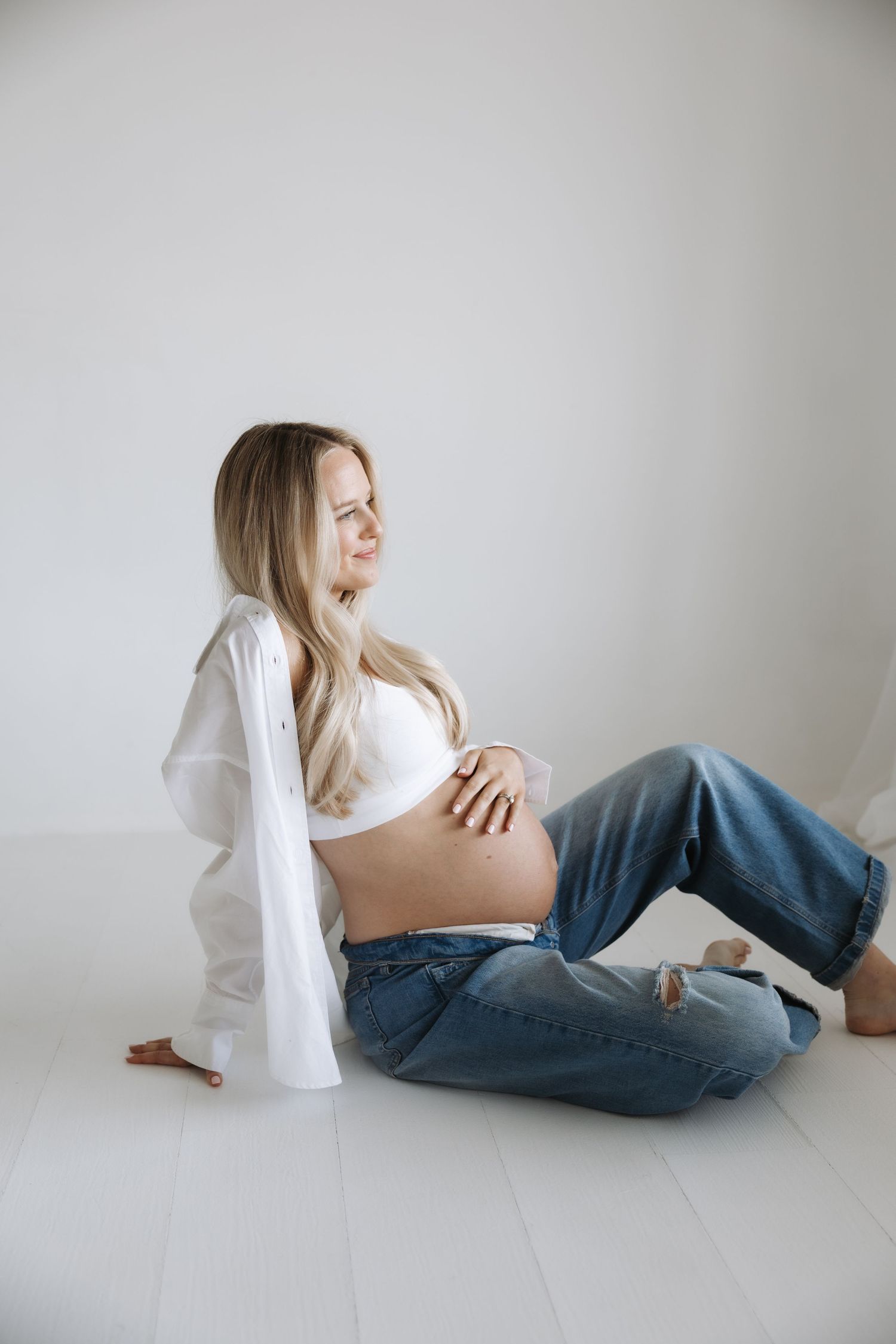 Pregnant woman in white crop top and jeans sitting on floor, gently cradling her baby bump in a bright studio.