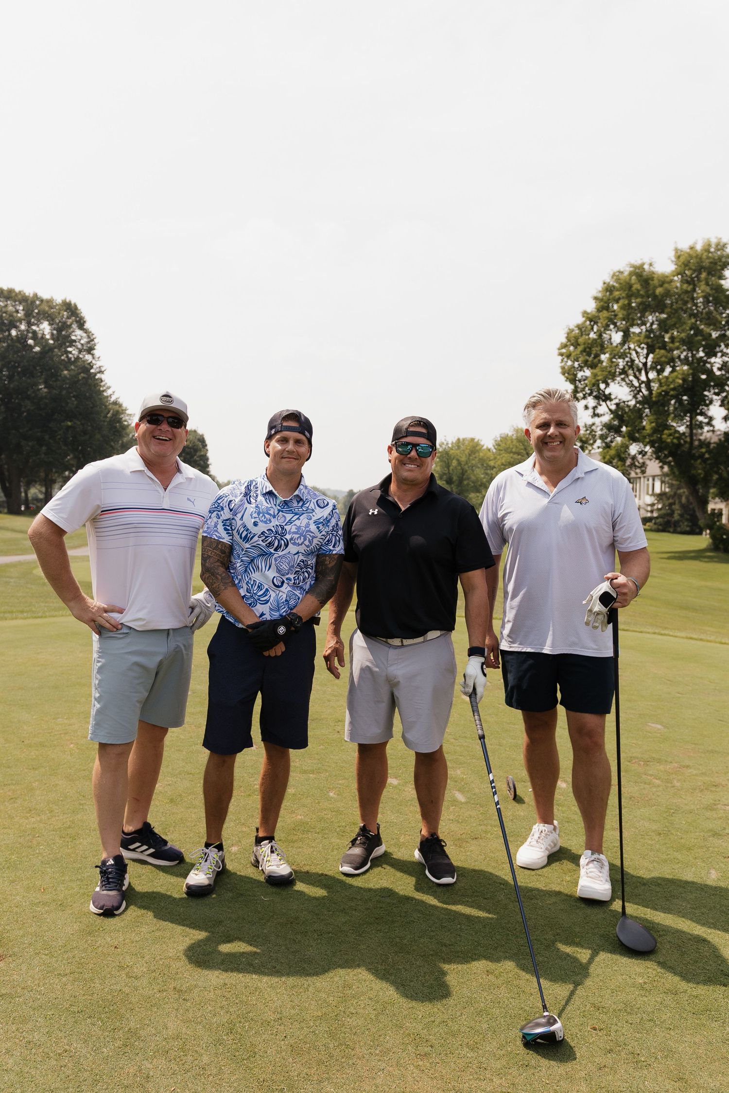 Four men posing together on a golf course on a sunny day, one holding a golf club.