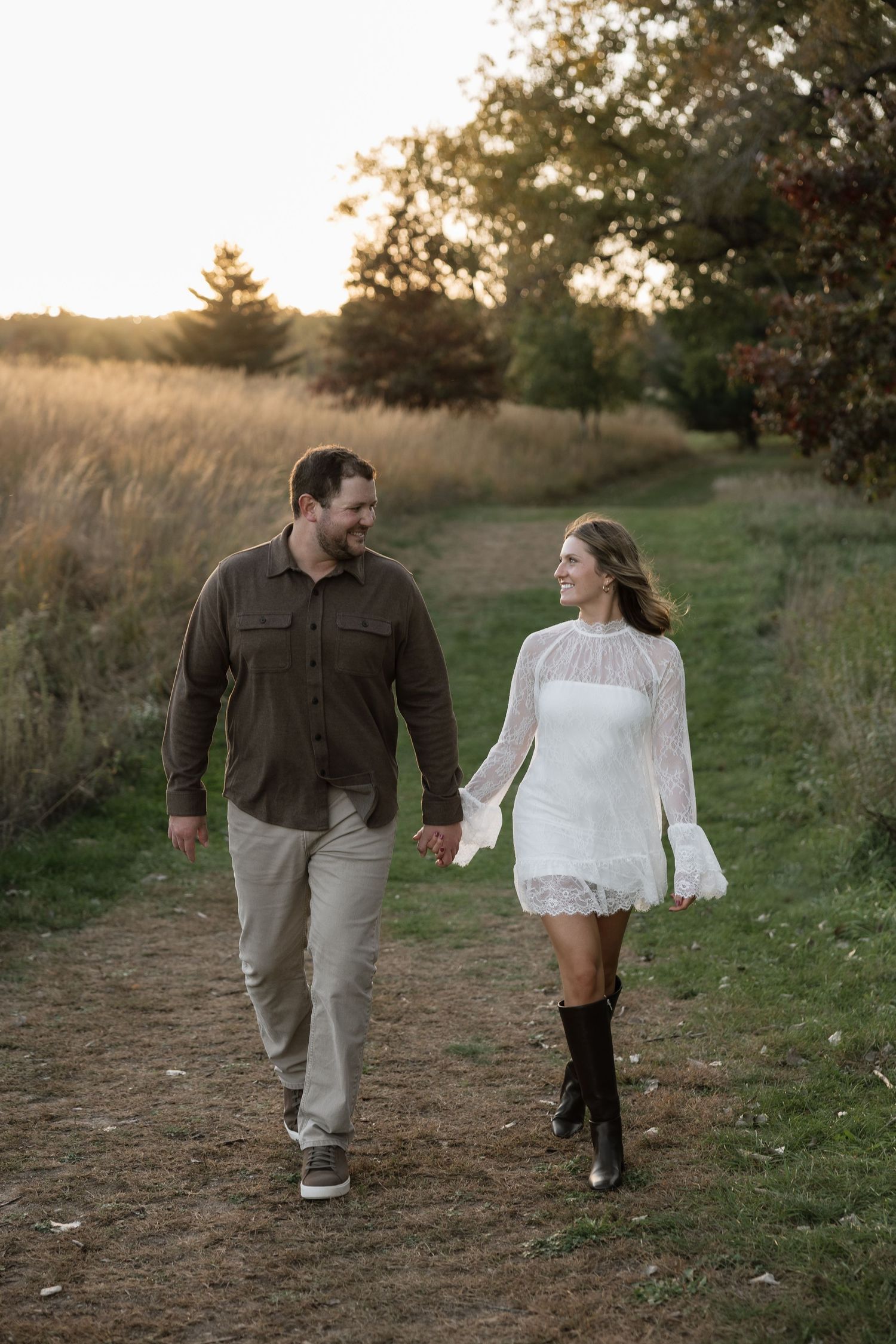 Happy couple walking hand in hand on a dirt path through a scenic field at golden hour.