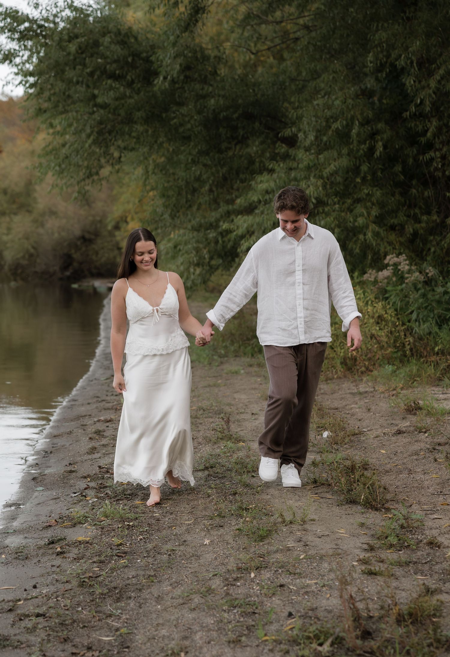 Couple holding hands walking along a sandy riverbank in white outfits, surrounded by lush green trees.