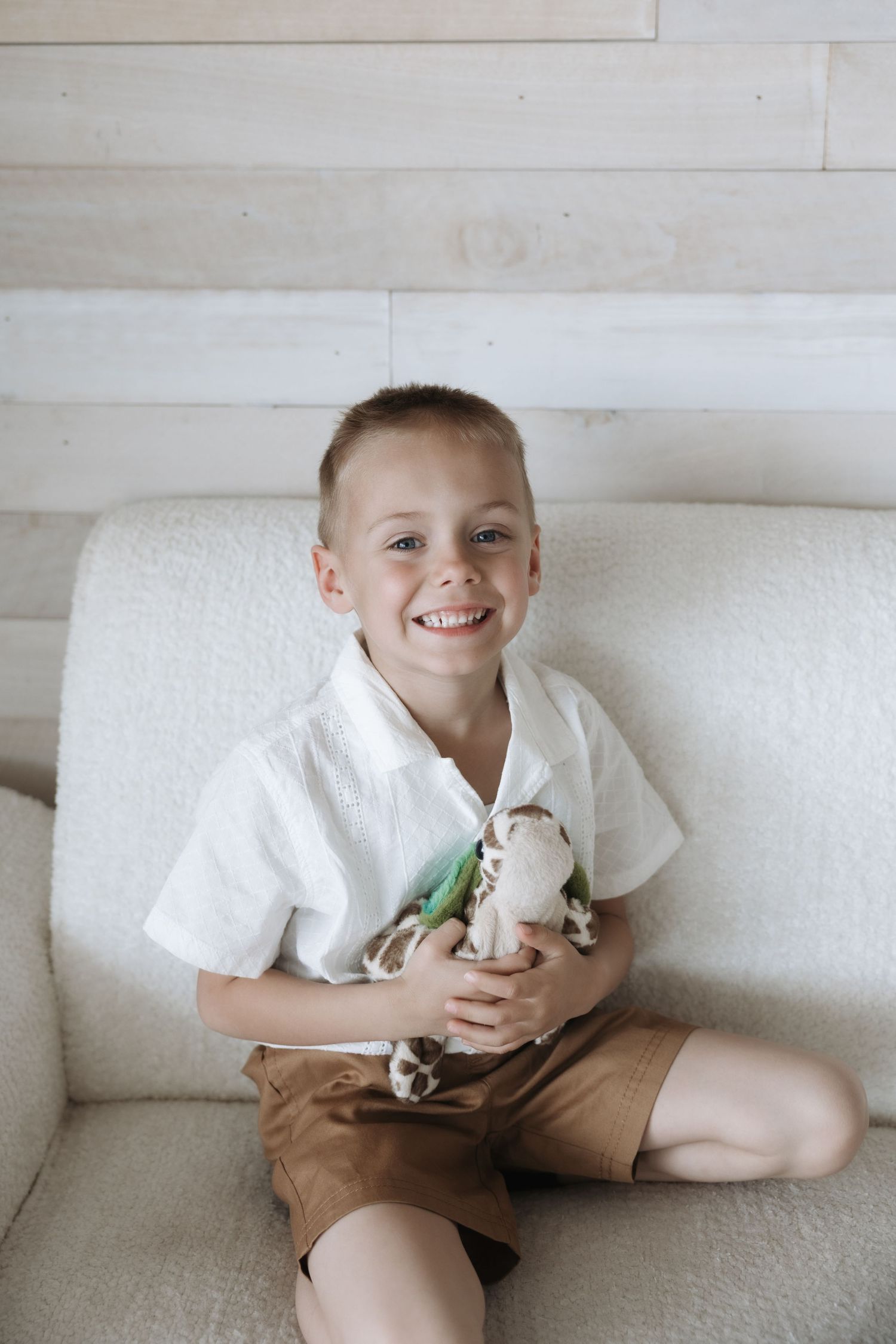 Smiling young boy in white shirt and tan shorts sitting on a chair holding stuffed toys.