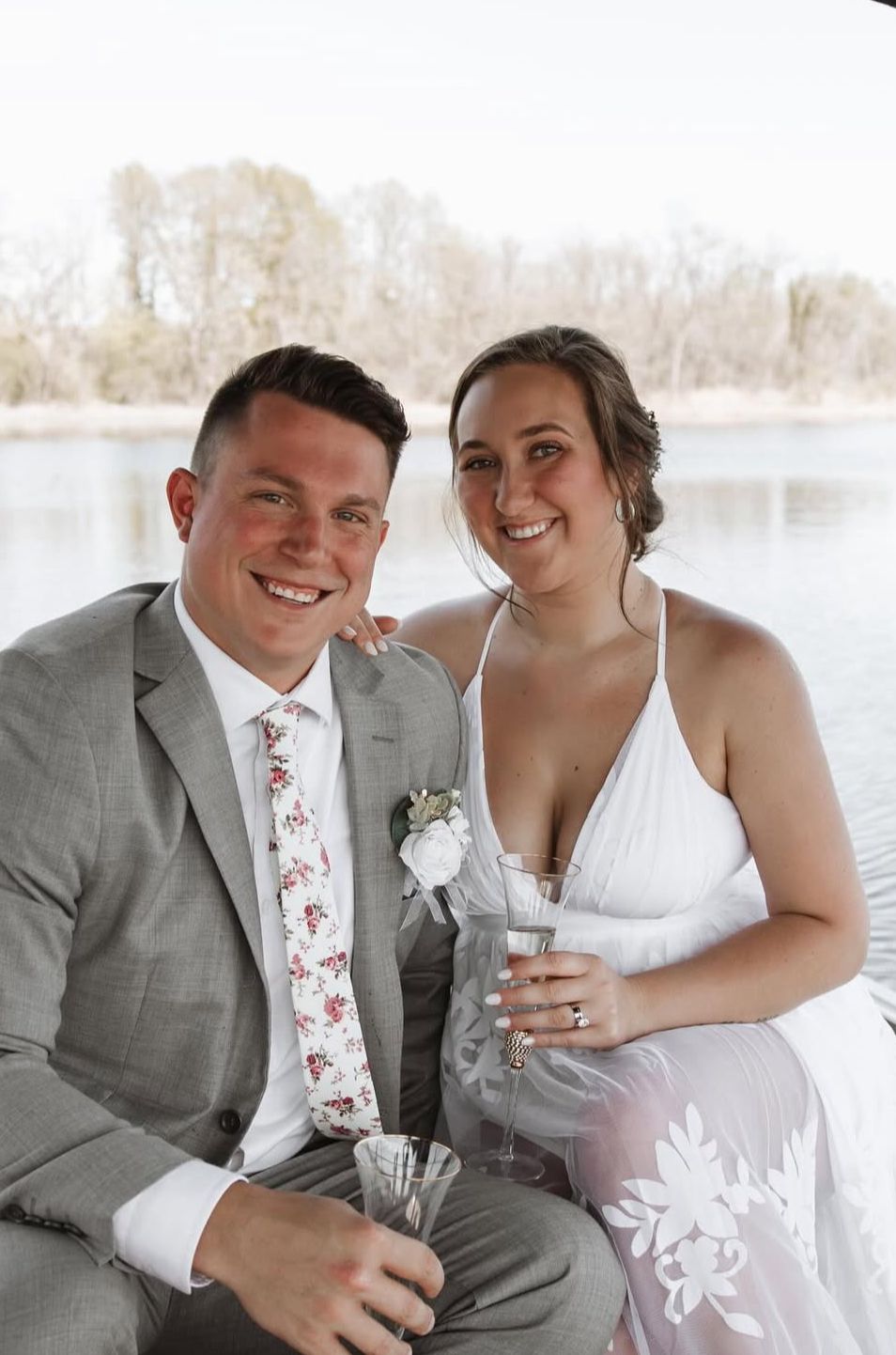 Happy couple in wedding attire sitting by a lake, groom in gray suit with floral tie, bride in white halter dress holding champagne.