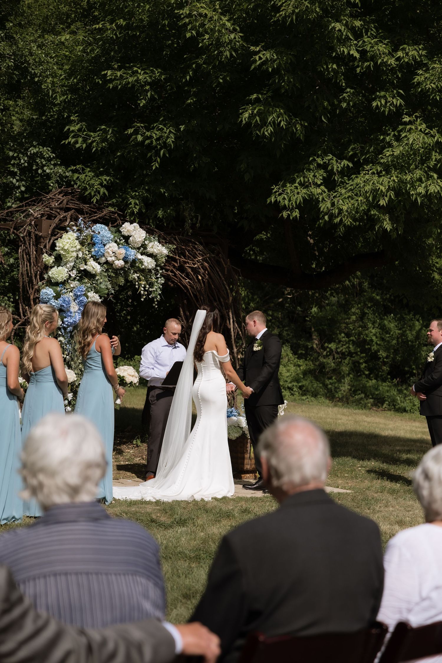 Bride and groom exchange vows at an outdoor ceremony beneath a floral arch with blue and white hydrangeas.