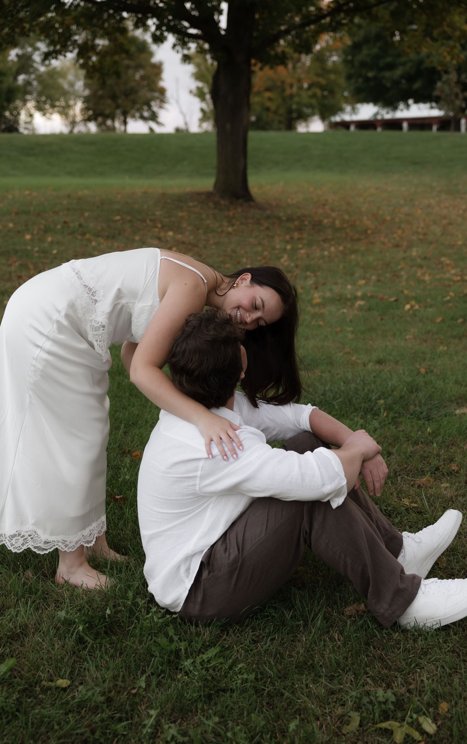 Loving couple in a park, woman in white dress leaning over seated man in white shirt on green grass.